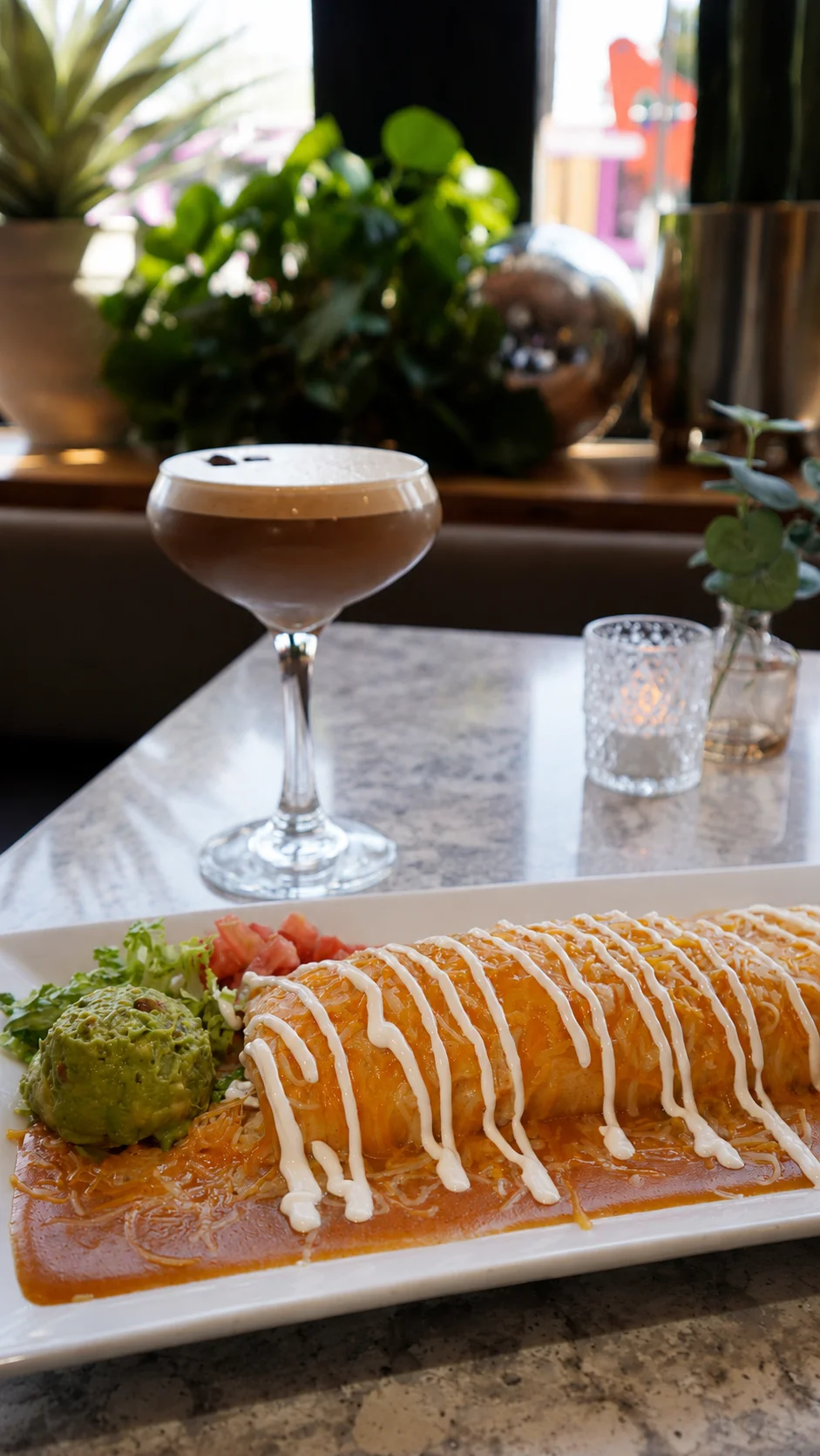 A gourmet plate with an enchilada topped with cheese, alongside guacamole and fresh ingredients, paired with a cocktail.