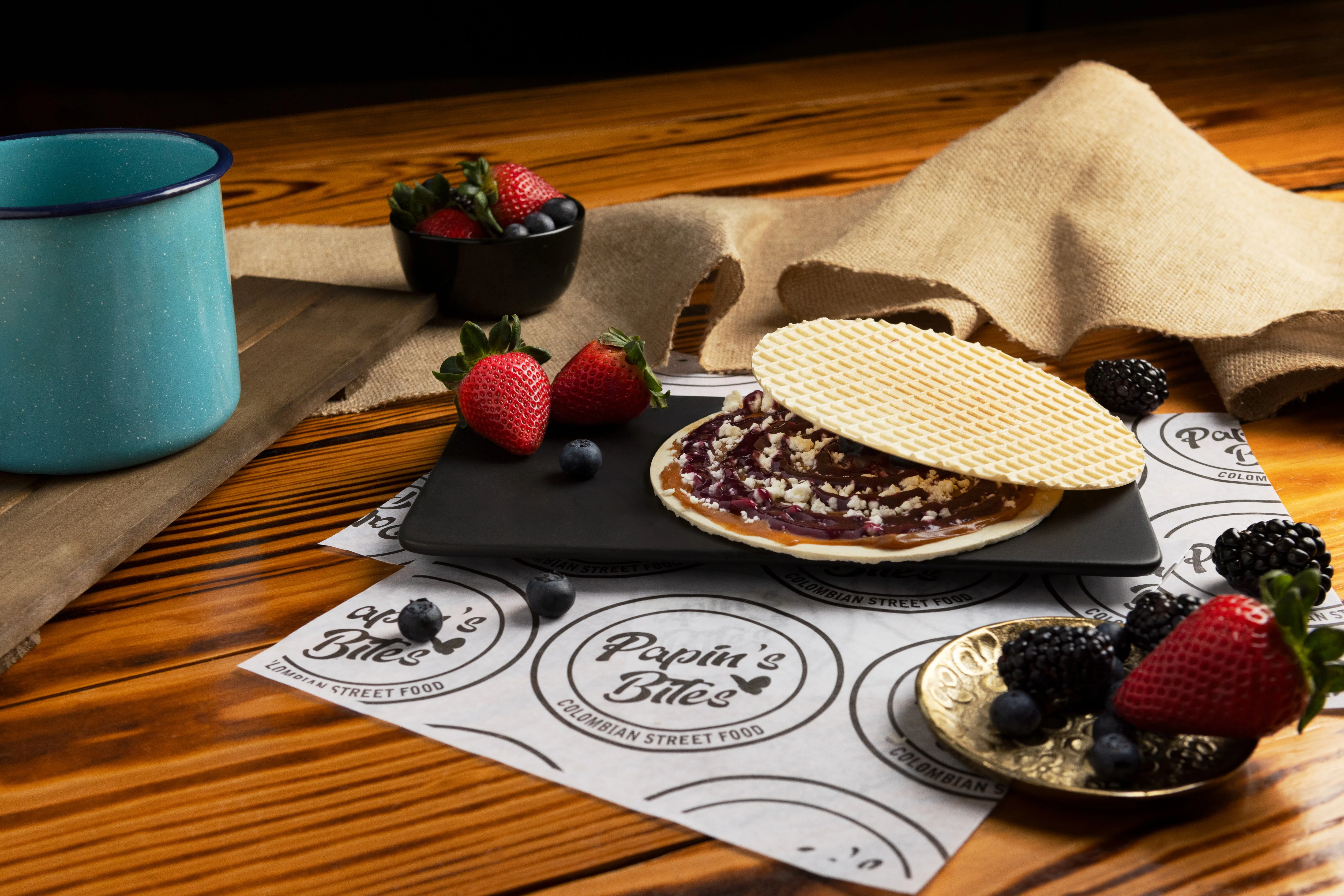 A flat dessert composed of a sweet wafer topped with a spread and served with fresh berries on a black plate. A rustic mug is also included in the scene.