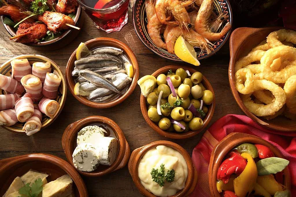 An assortment of tapas featuring shrimp, olives, bacon-wrapped items, calamari, and various dips, arranged in small bowls.