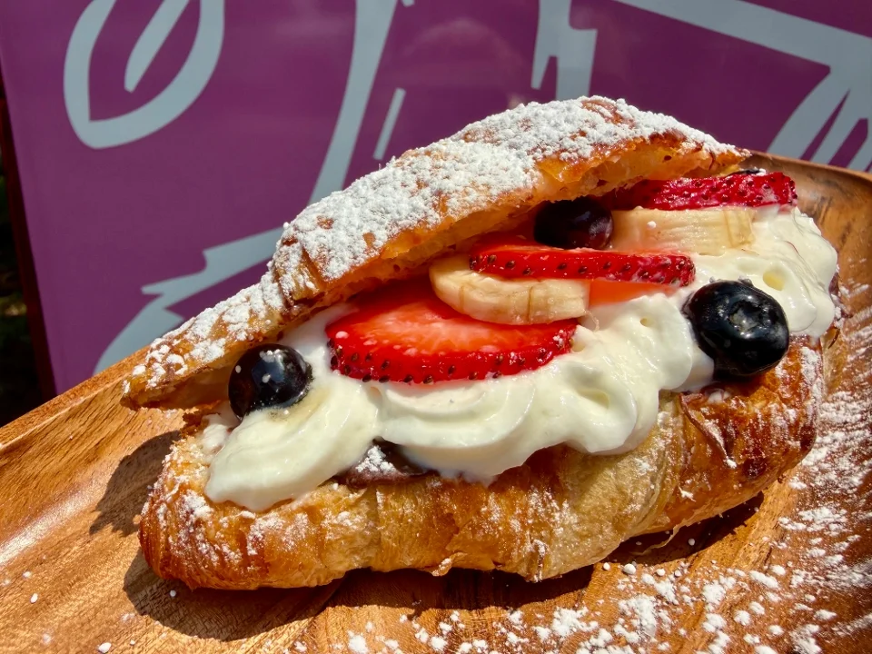 A croissant sandwich filled with cream, bananas, strawberries, and blueberries, dusted with powdered sugar on a wooden plate.