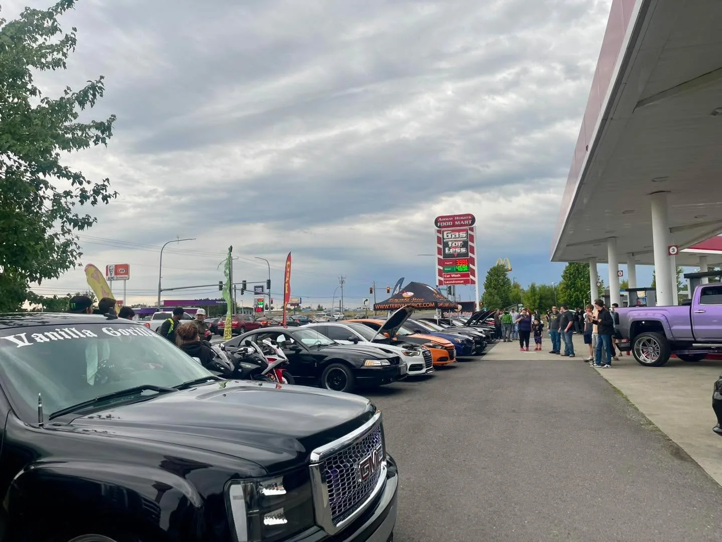 A gathering of various cars parked alongside a gas station with people admiring them. The sky is overcast.