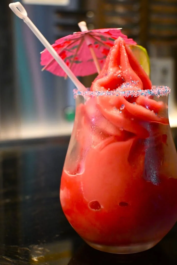 A vibrant frozen drink topped with a swirl of slushy texture, garnished with a pink umbrella and a slice of lime. The glass is rimmed with colorful sugar.