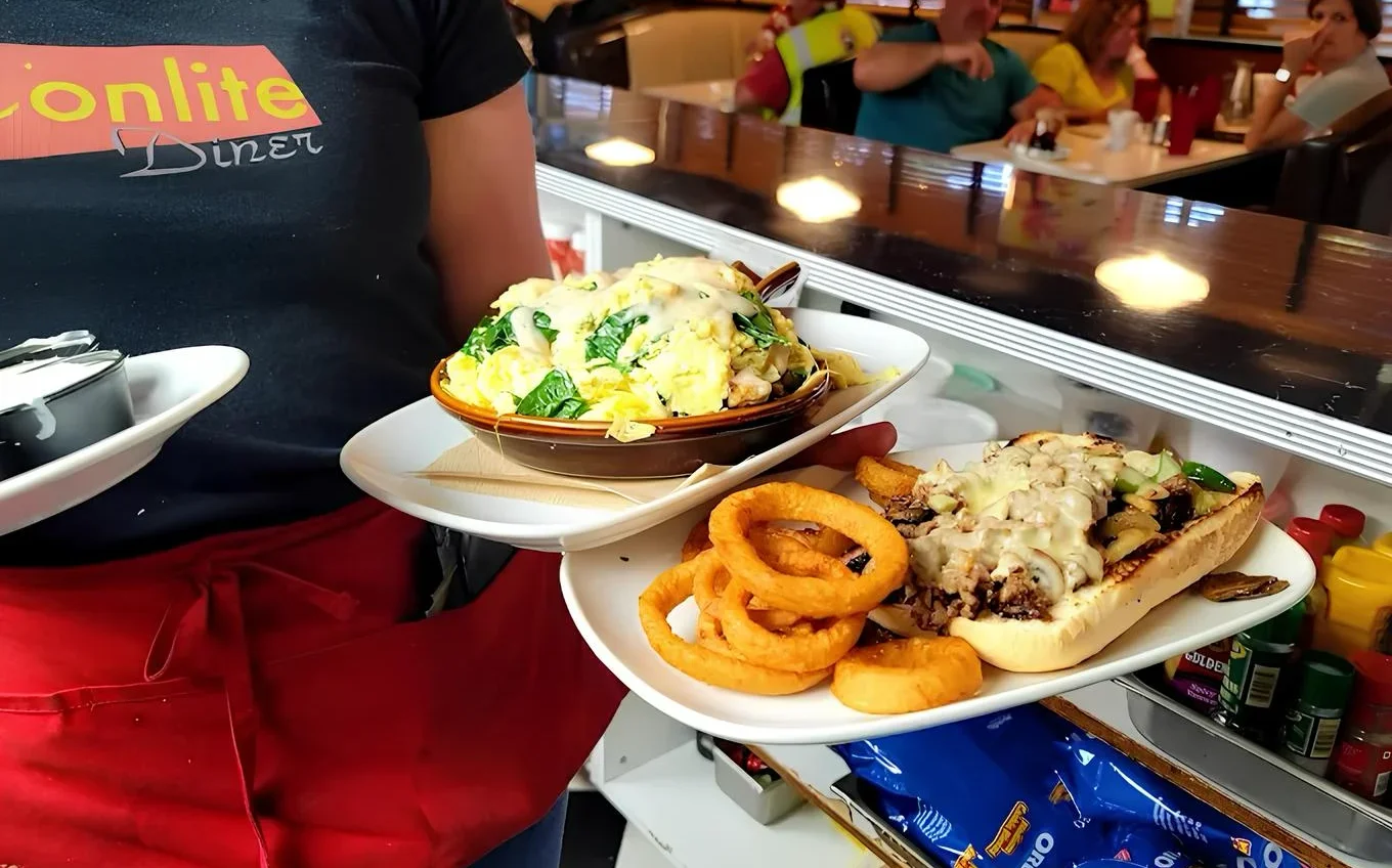 Server carrying signature diner meals, including a cheesesteak sandwich with onion rings and a baked entrée, at Moonlite Diner.