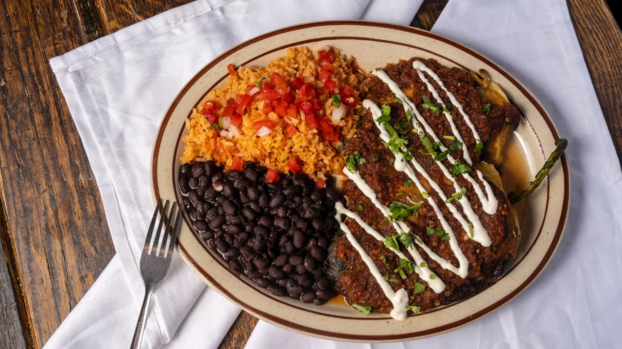 A plate featuring a serving of black beans, rice with diced tomatoes, and two pieces of meat topped with sour cream and herbs.