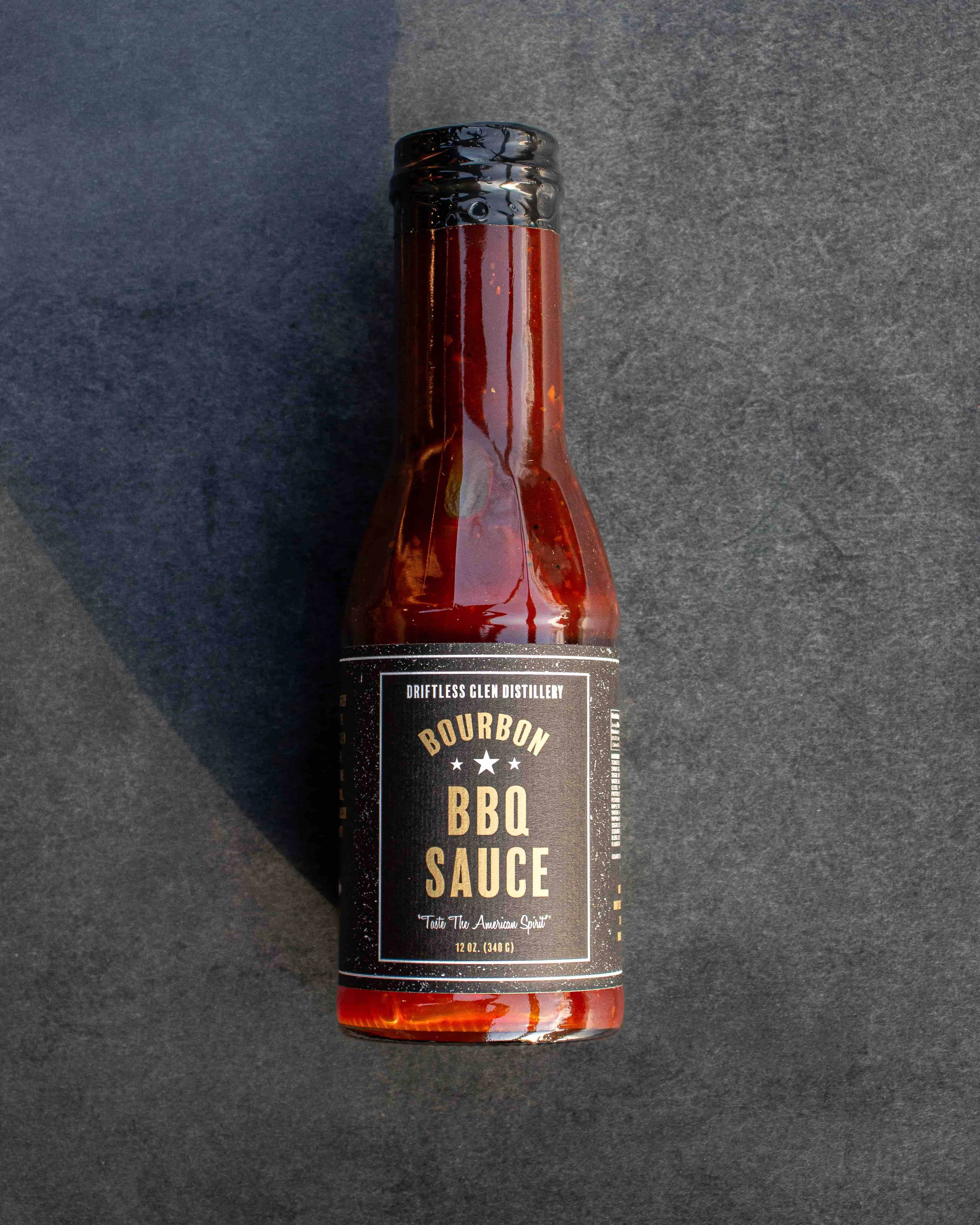 A bottle of Driftless Glen's Bourbon BBQ Sauce sits on a dark tabletop with light shining on it.