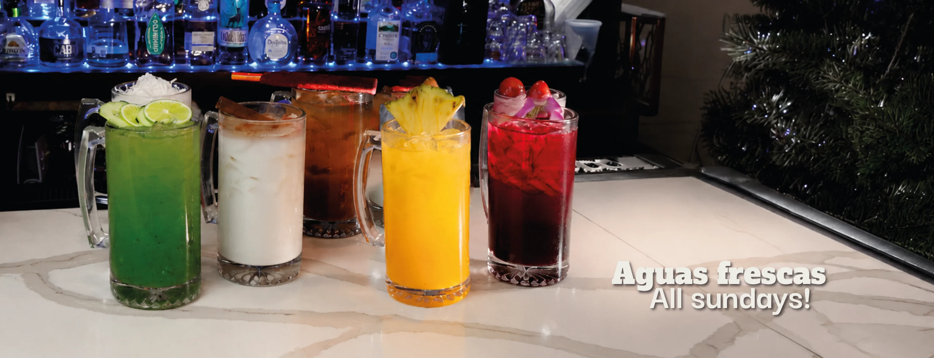 Five colorful aguas frescas drinks on a bar counter with festive decor.