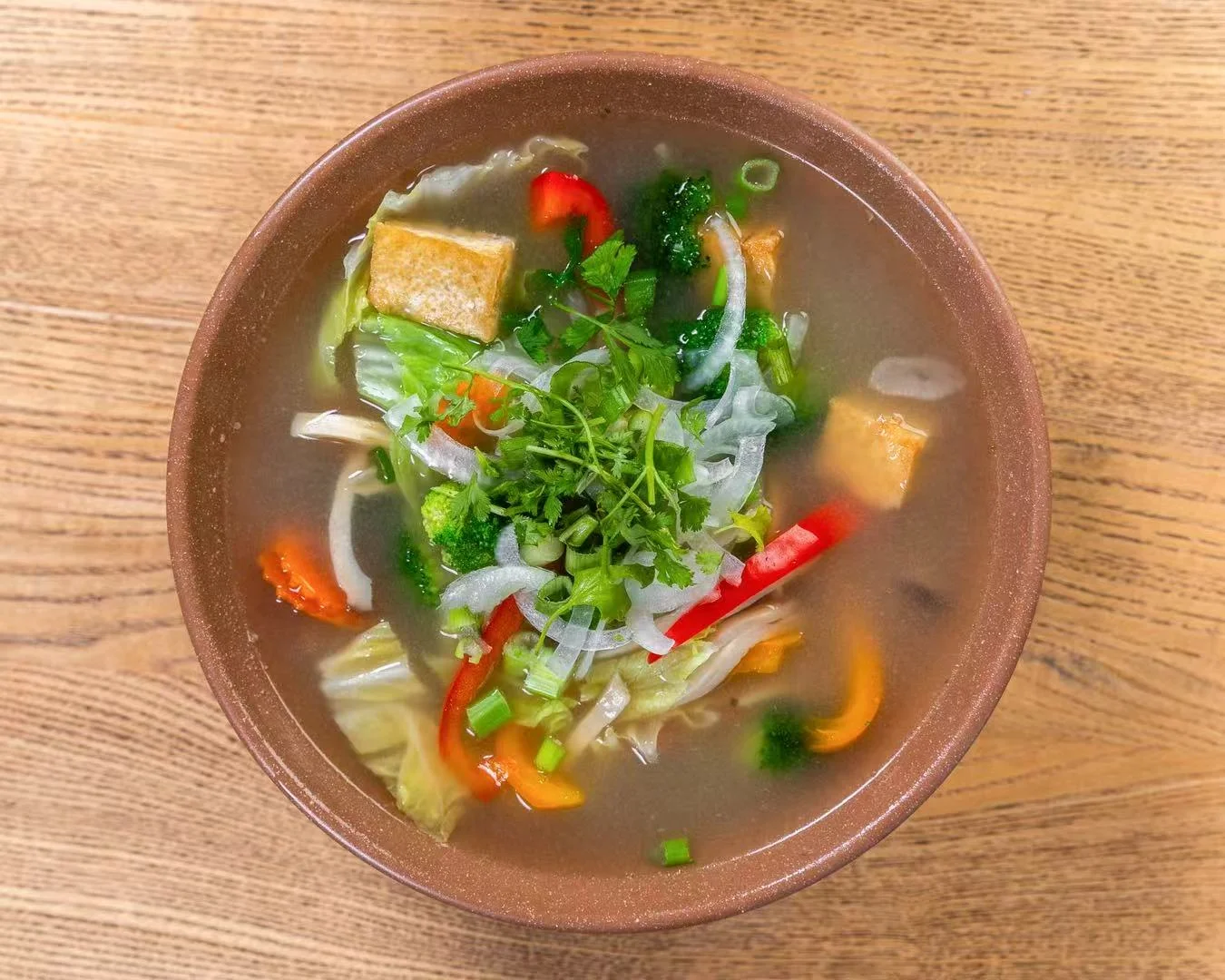 Bowl of vegetable soup with tofu and fresh herbs on a wooden table.