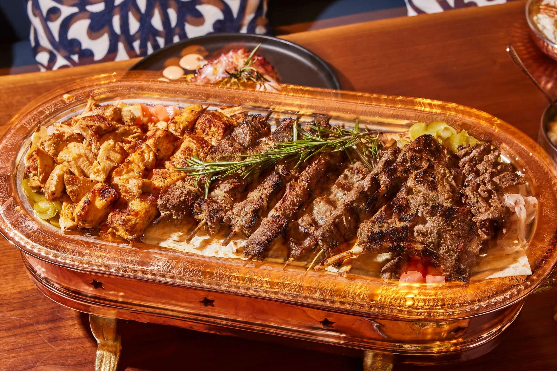 A wooden platter with assorted grilled skewers, garnished with rosemary. Behind is a black plate with dipping sauces, creating a warm, appetizing scene.
