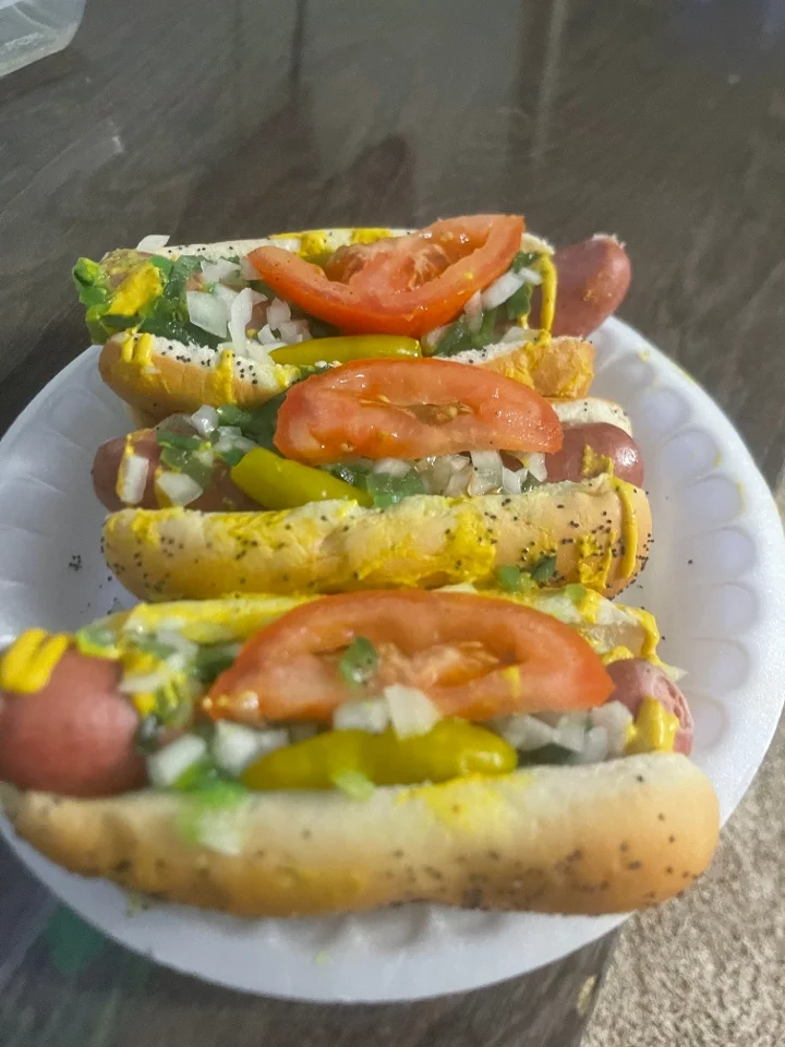 A plate of hot dogs topped with mustard, tomatoes, onions, and jalapeños, served on a white paper plate.