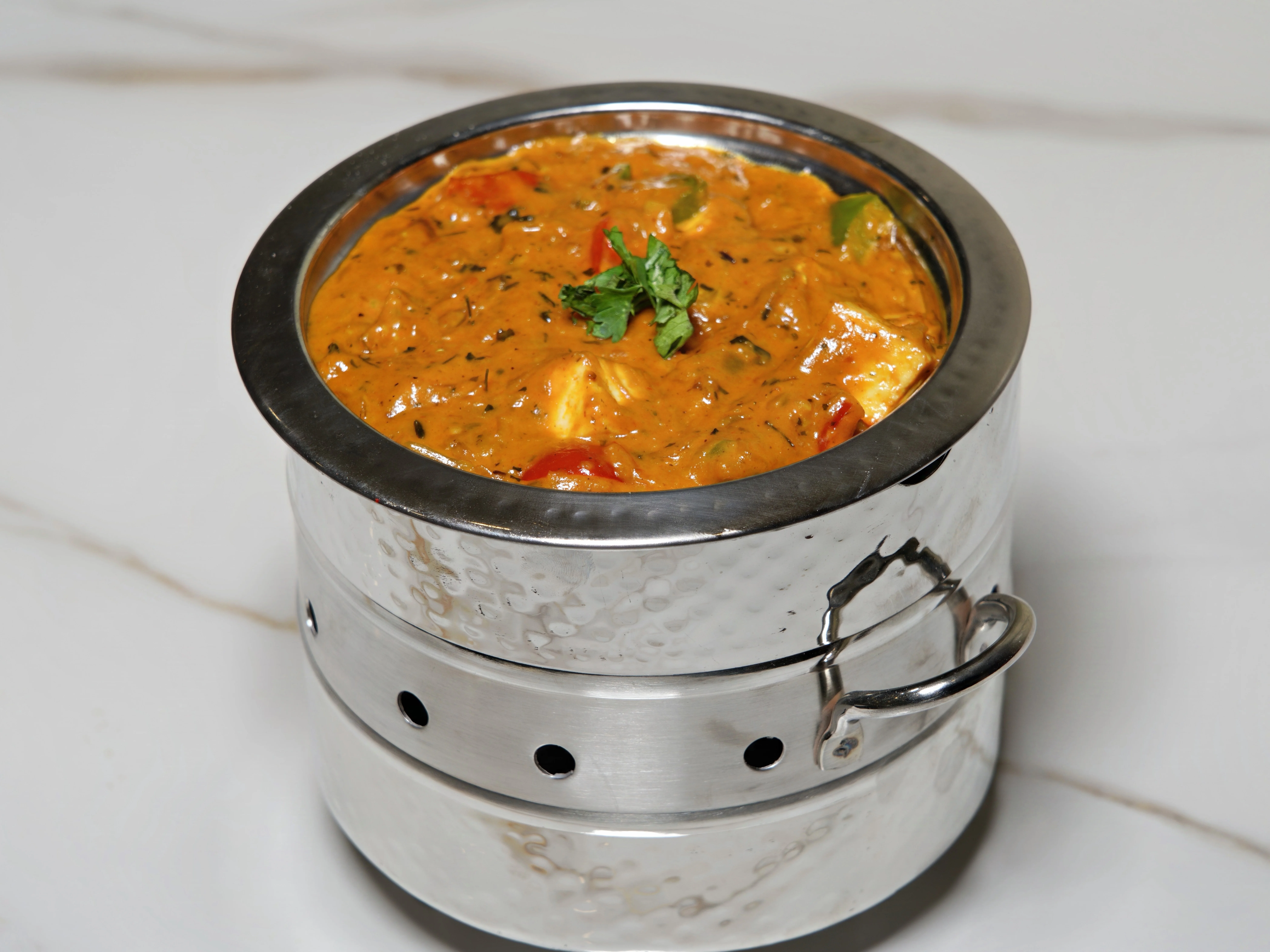 A serving of a rich and creamy curry dish, garnished with herbs, served in a traditional metal container.