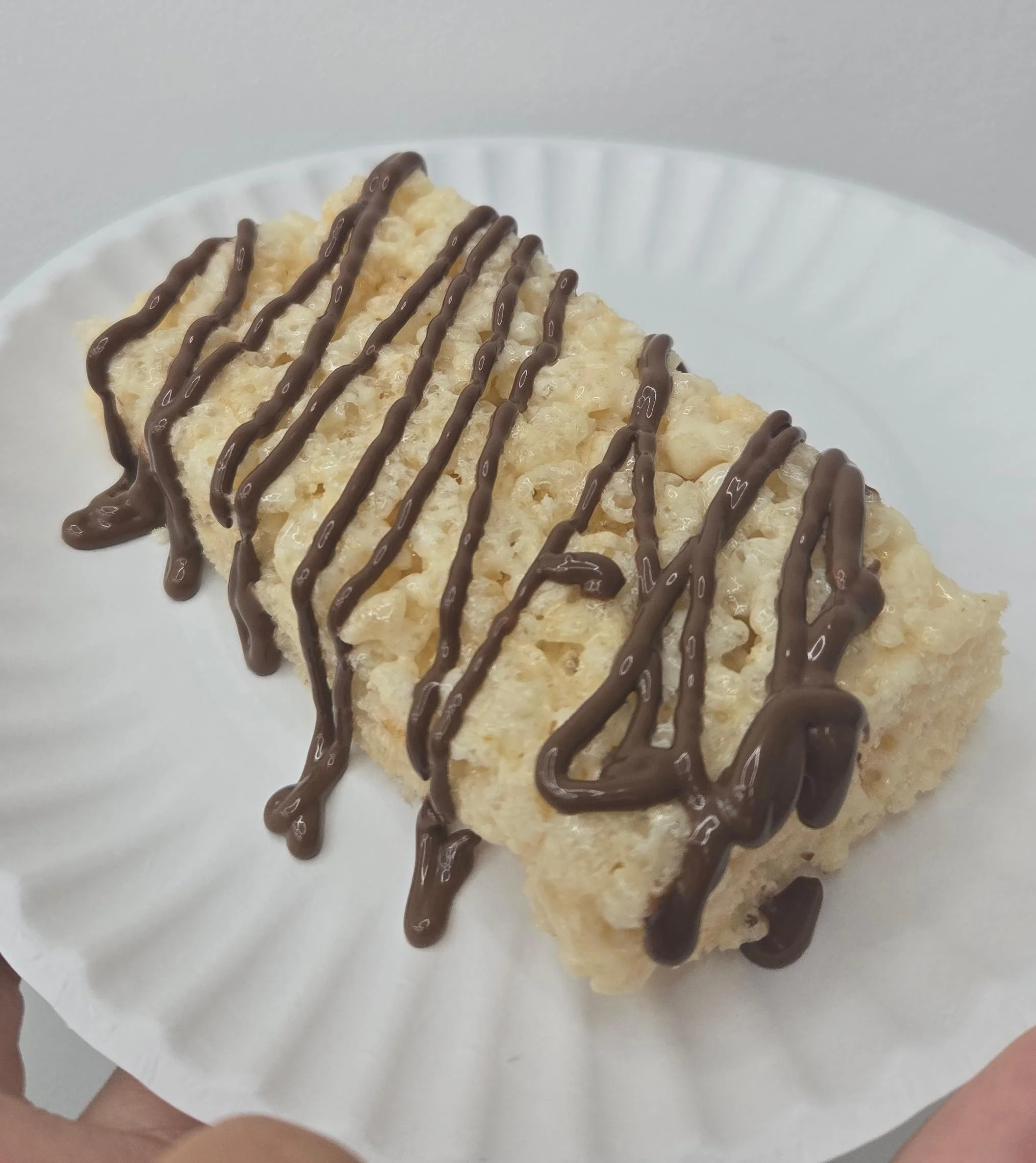 A rectangular rice krispie treat drizzled with chocolate, placed on a white paper plate.