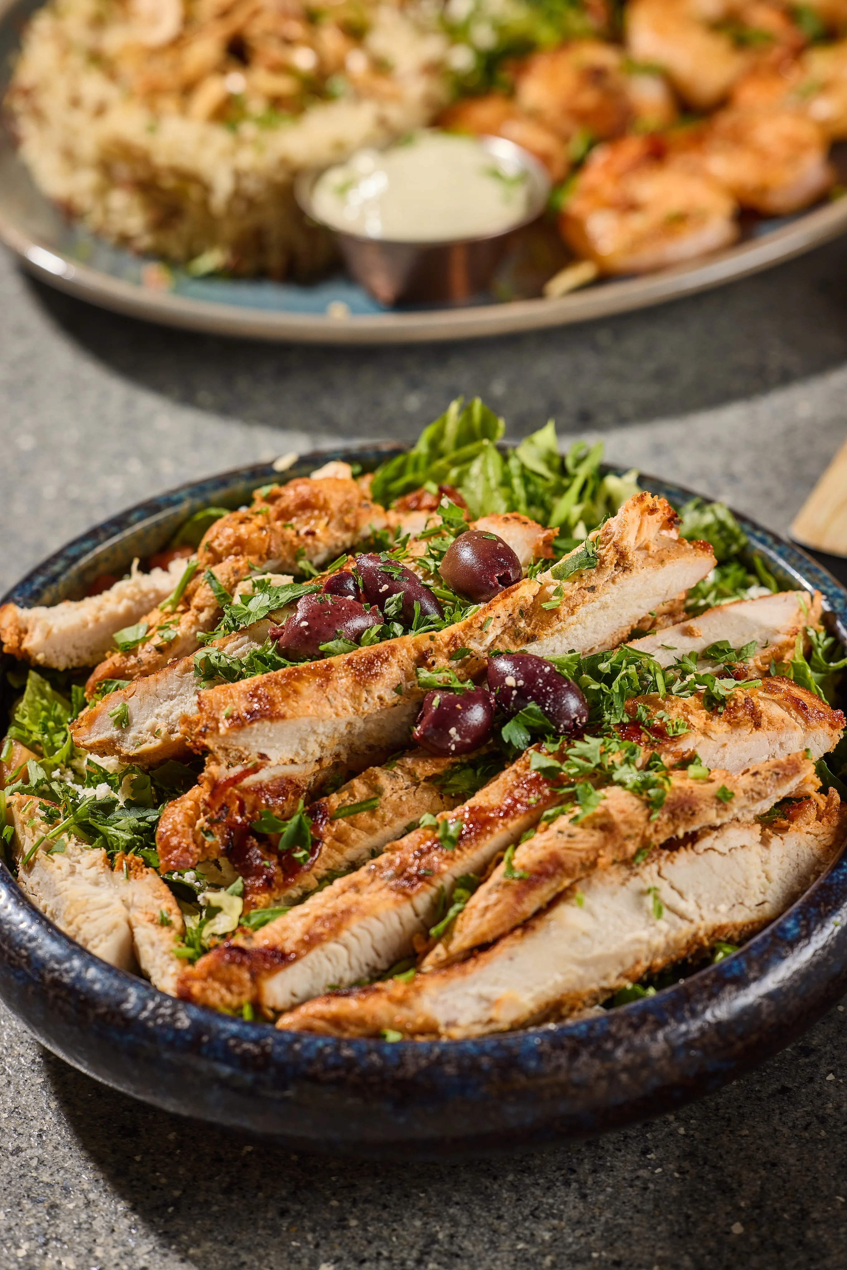 Grilled chicken strips on a bed of fresh green lettuce, garnished with purple olives and herbs, served in a dark blue bowl, creating a vibrant and appetizing display.