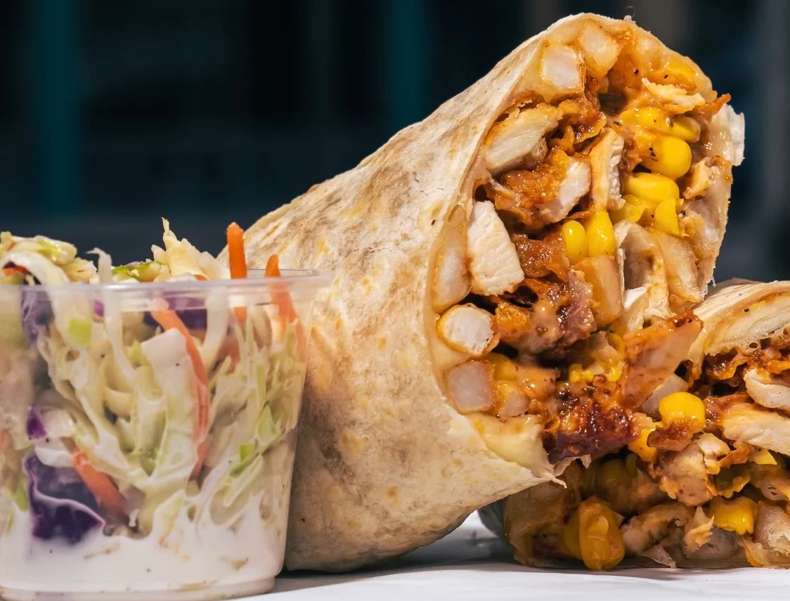 A close-up of a wrapped burrito with chicken, corn, and other fillings, accompanied by a side of coleslaw in a cup.