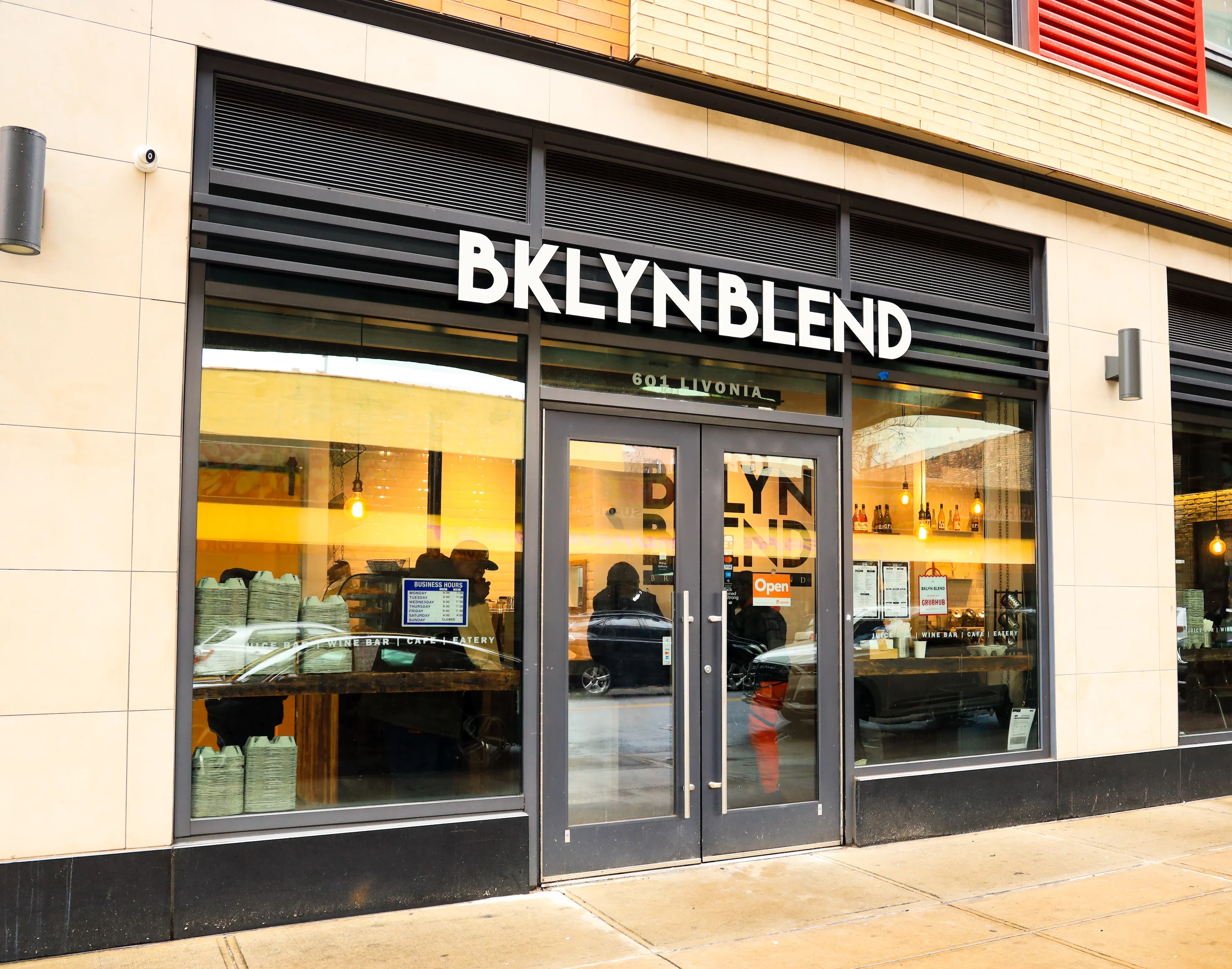 Front of building with BKLYN BLEND in large white letters above door, double glass doors with large glass windows, black door and window frames, people are inside, yellow hanging lights light up the interior