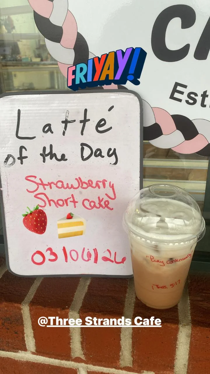 A sign advertising the latte of the day, featuring a strawberry shortcake flavor, next to an iced latte in a clear cup.