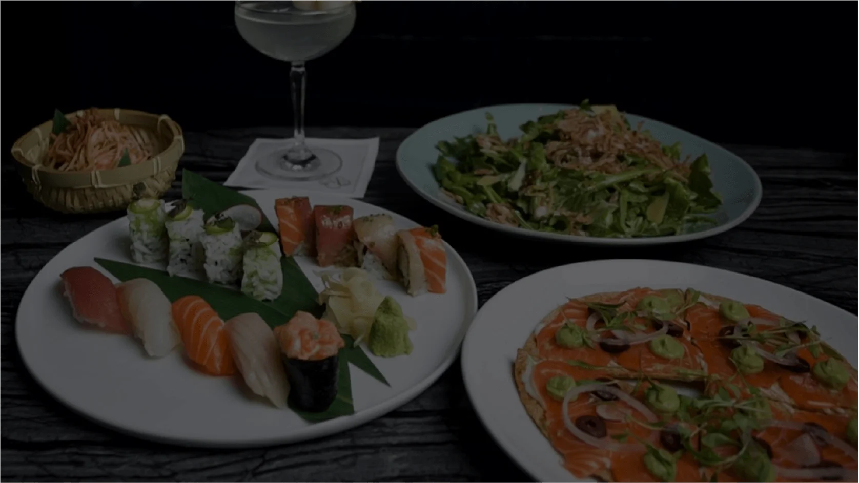 A gourmet restaurant table setting featuring a large round plate of assorted sushi, including nigiri and uramaki rolls served with wasabi and ginger, alongside a plate of salmon carpaccio flatbread topped with avocado crema, a fresh green salad with fried shallots, a small bamboo basket of shredded vegetables, and a coupe cocktail garnished with lychees on a dark, textured stone surface.