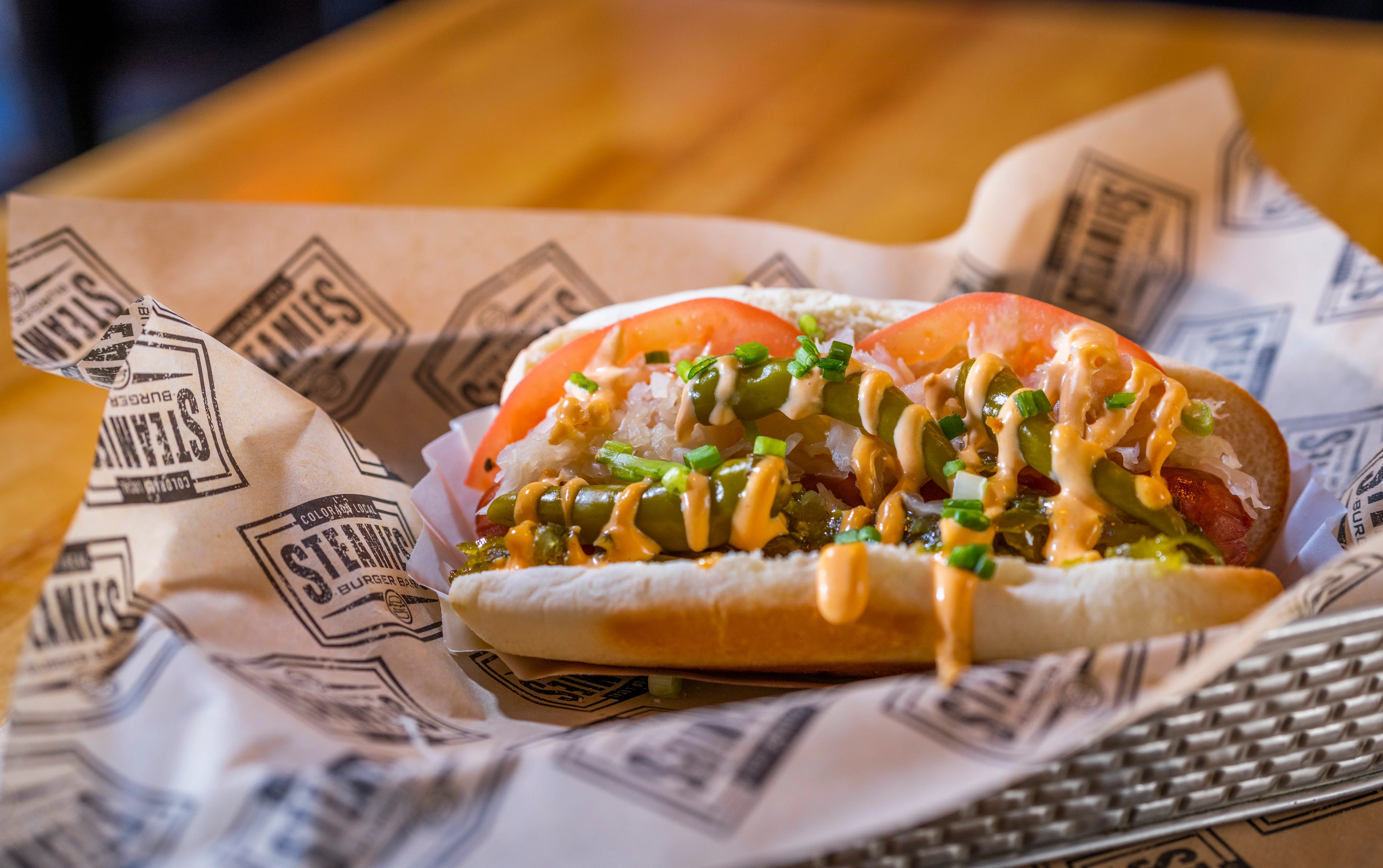 A hot dog filled with toppings including tomatoes, green onions, and a drizzle of sauce, served in a paper-lined basket.