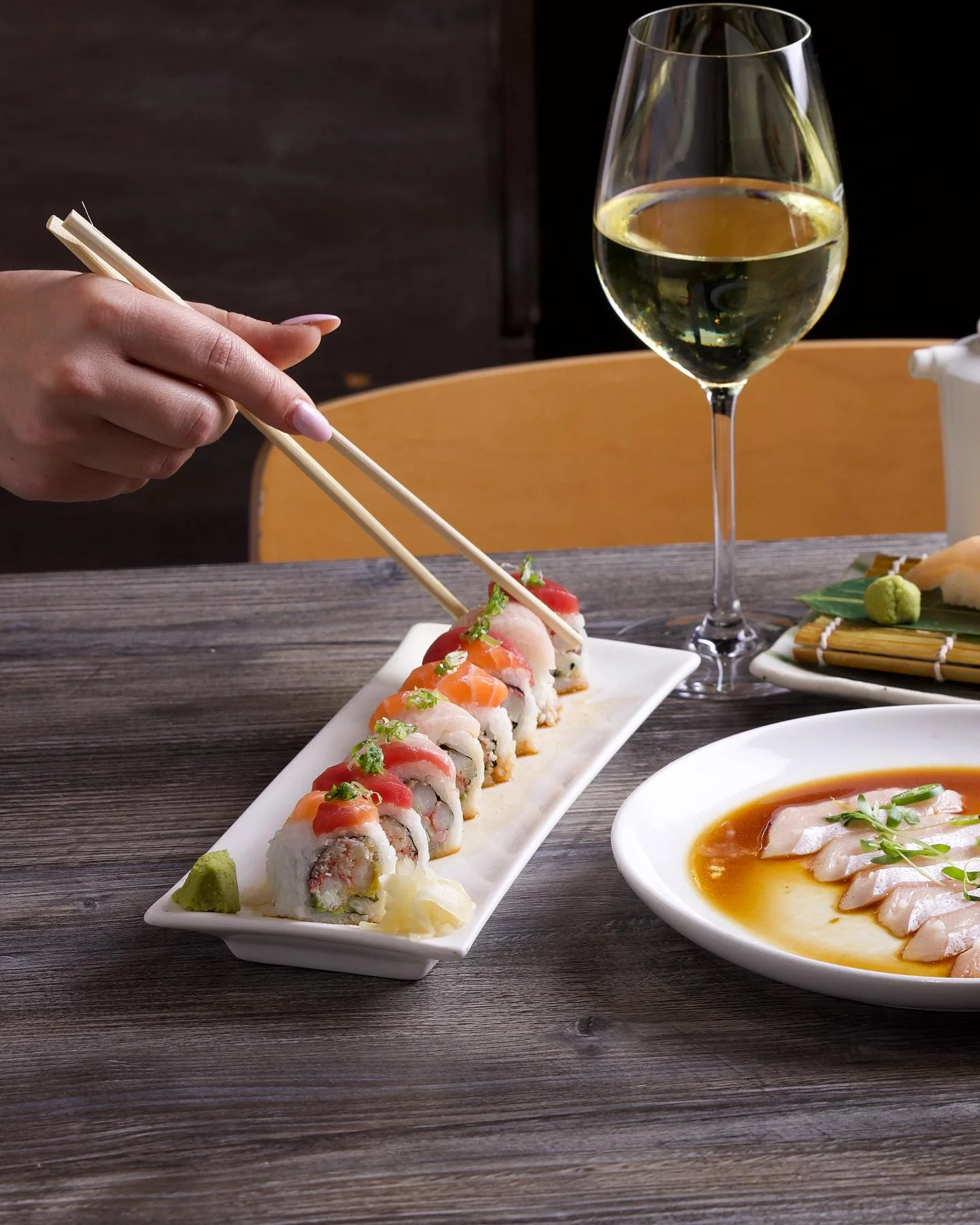 A variety of fresh sushi rolls topped with tuna and salmon being picked up with chopsticks, served on a wooden table alongside a glass of white wine and thinly sliced yellowtail sashimi.