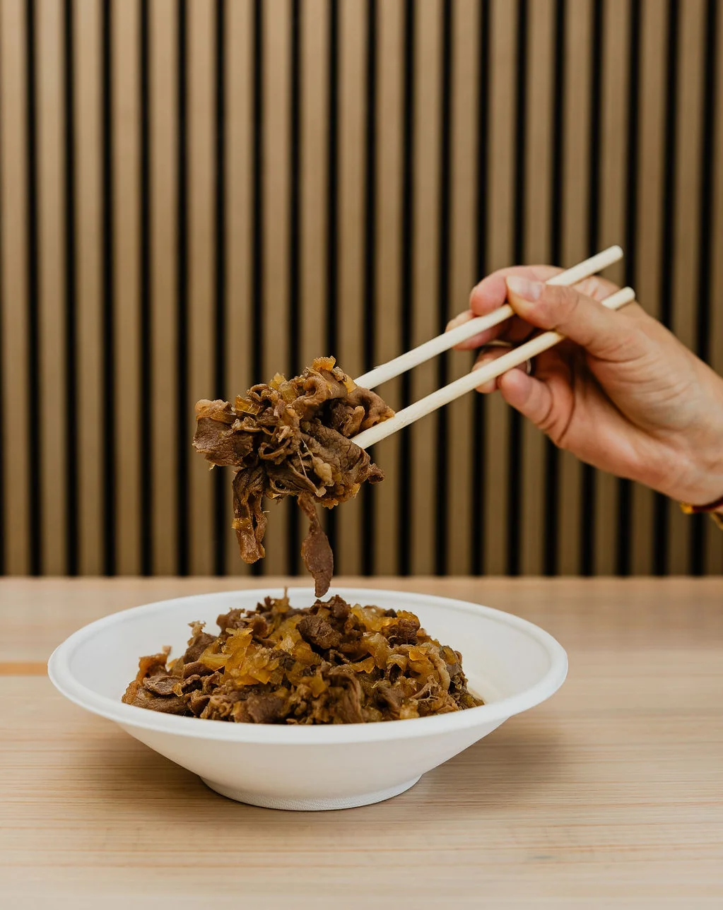 a hand holding up a portion of thinly sliced gyudon beef with chopsticks