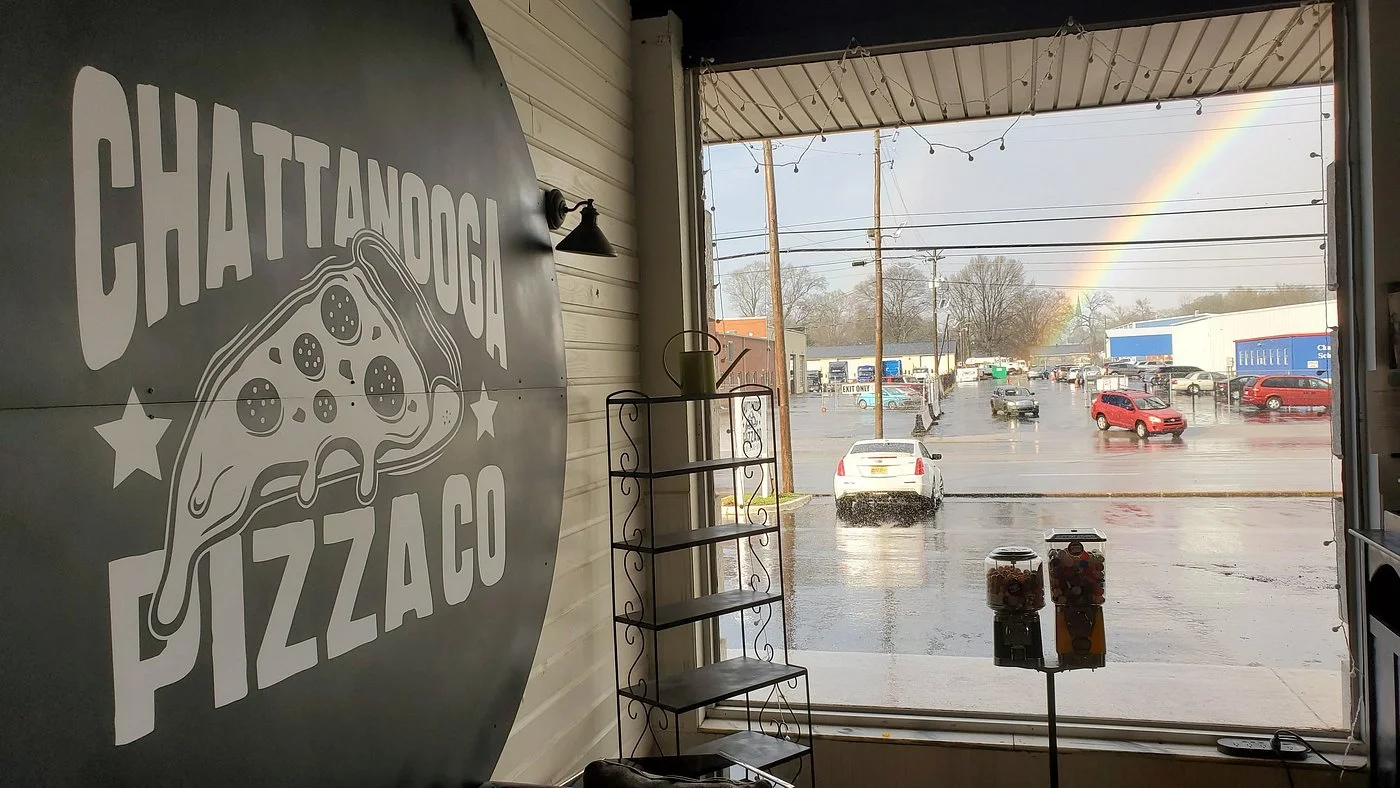 A view of a restaurant interior featuring a large pizza-themed sign, with rain outside and a rainbow visible through the window.