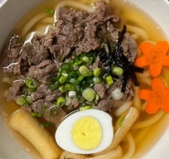 A bowl of noodle soup topped with sliced beef, green onions, seaweed, carrots, and a halved soft-boiled egg, served in a rich broth.
