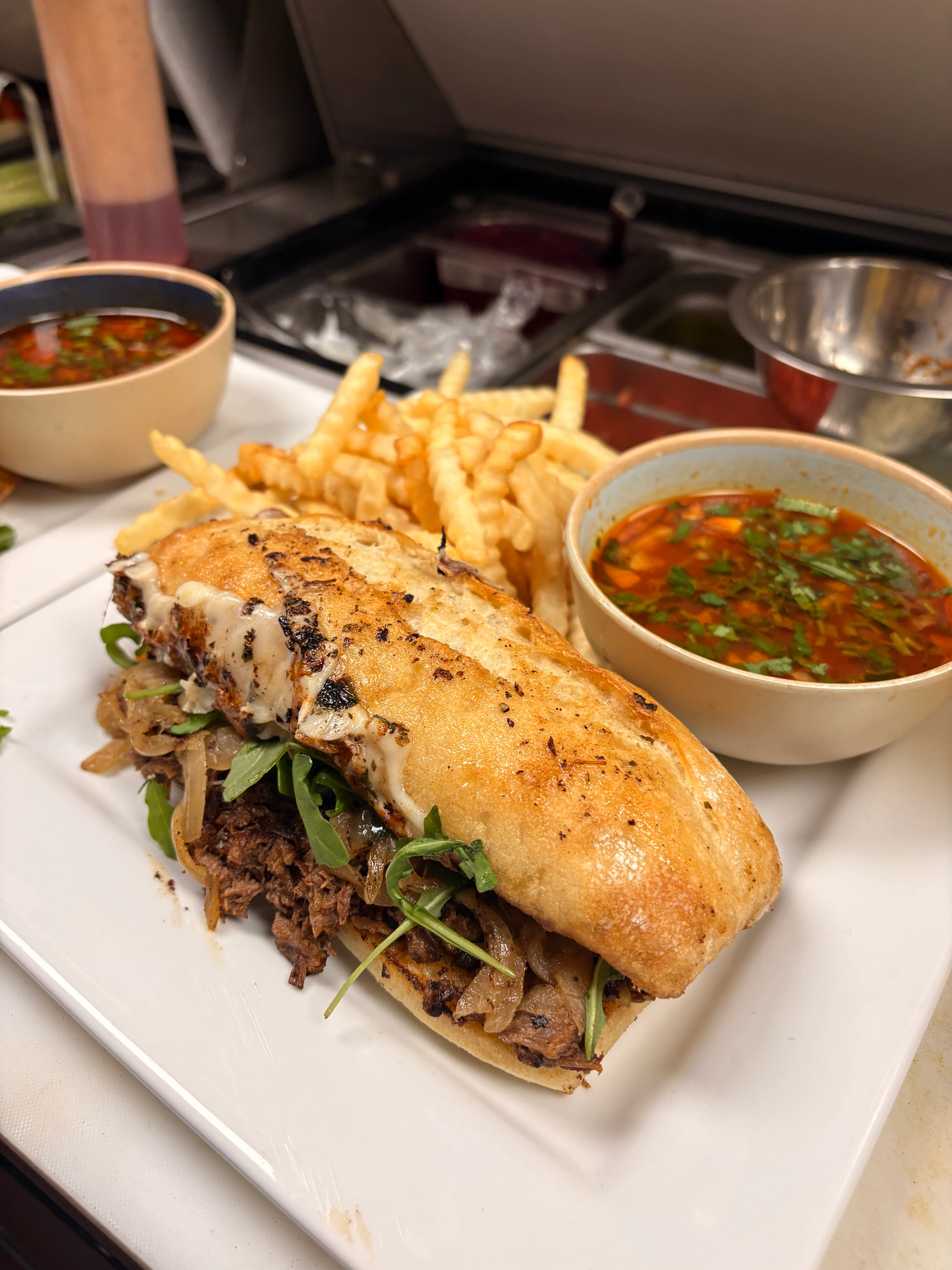 A savory sandwich filled with beef, grilled onions, and arugula, served with French fries and two bowls of dipping sauce.