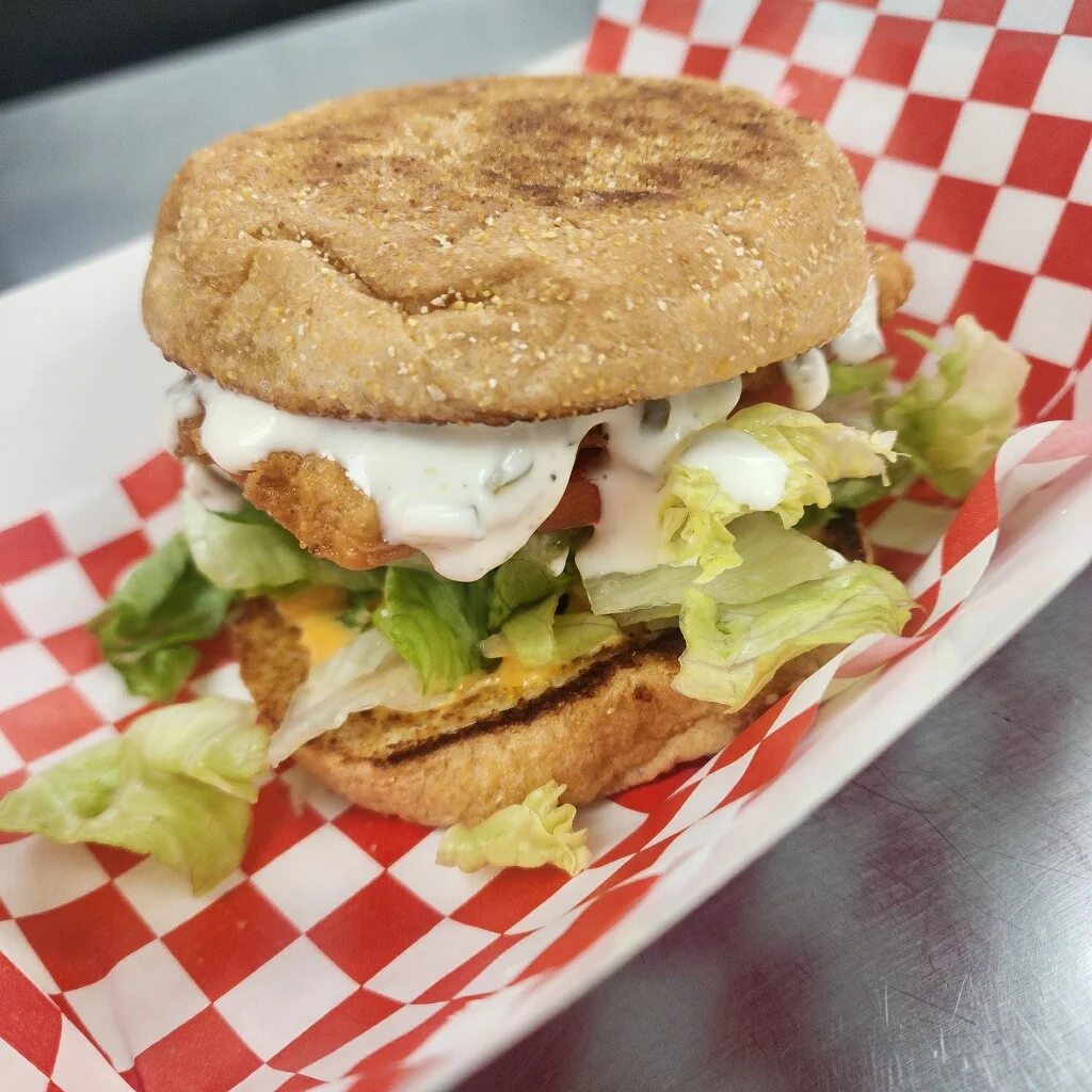 Close-up of a crispy chicken sandwich served on an English muffin, topped with shredded lettuce, tomato, and creamy ranch dressing, placed in a red-and-white checkered paper tray.