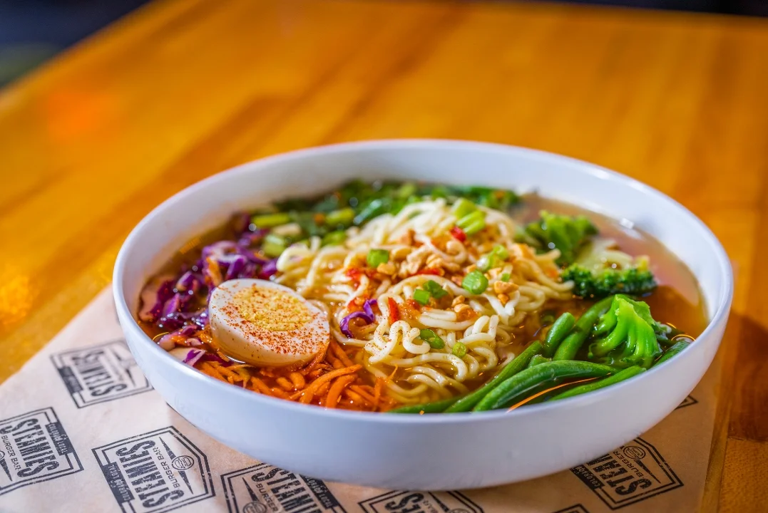 A bowl of ramen featuring noodles, colorful vegetables, and a soft-boiled egg, garnished with spices and herbs.