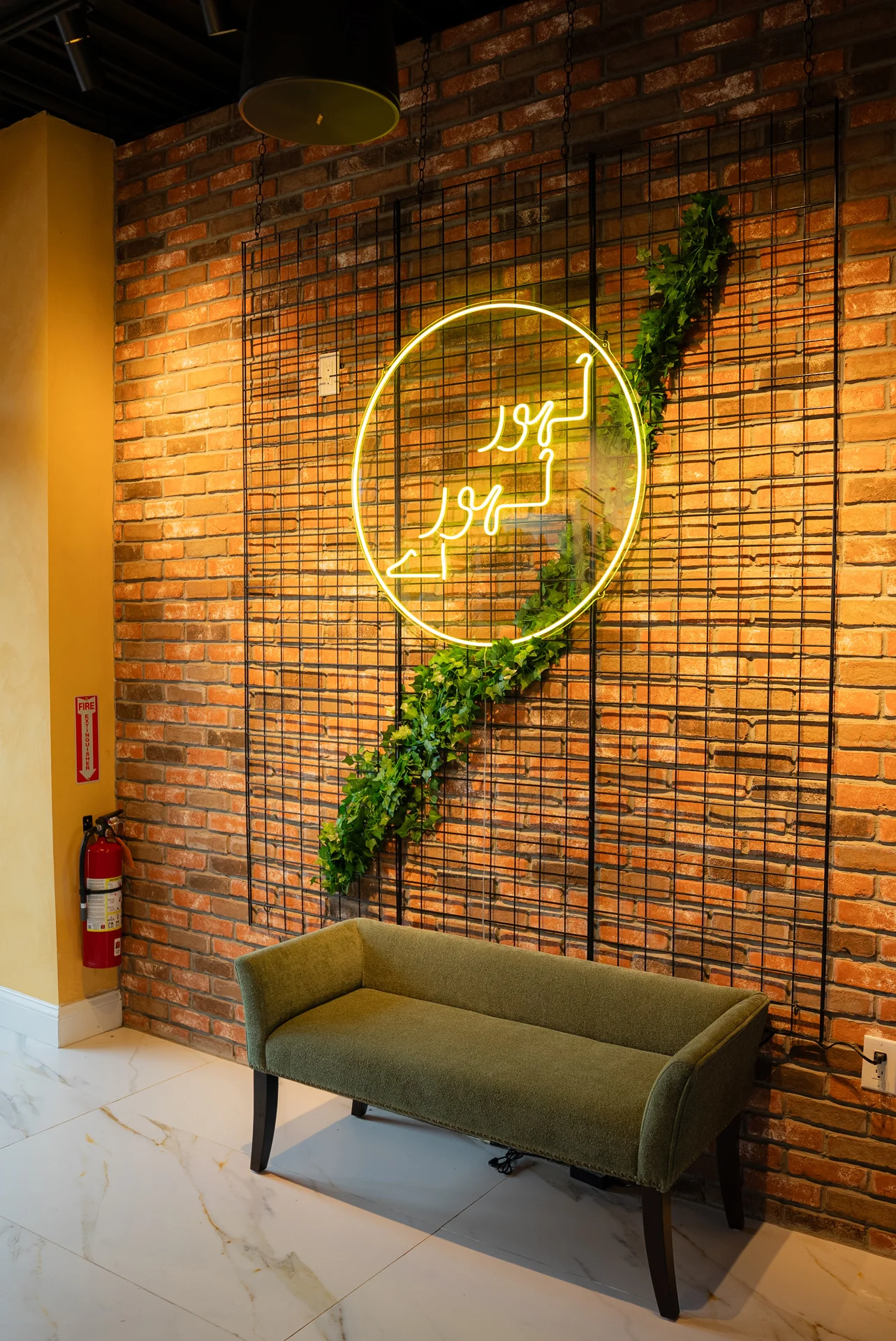 Bright neon sign above olive colored couch against a brick wall.