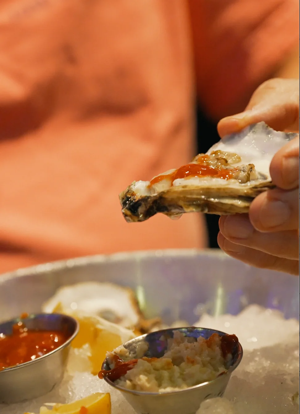A hand holding an oyster topped with sauce, with lemon and ice in the background.