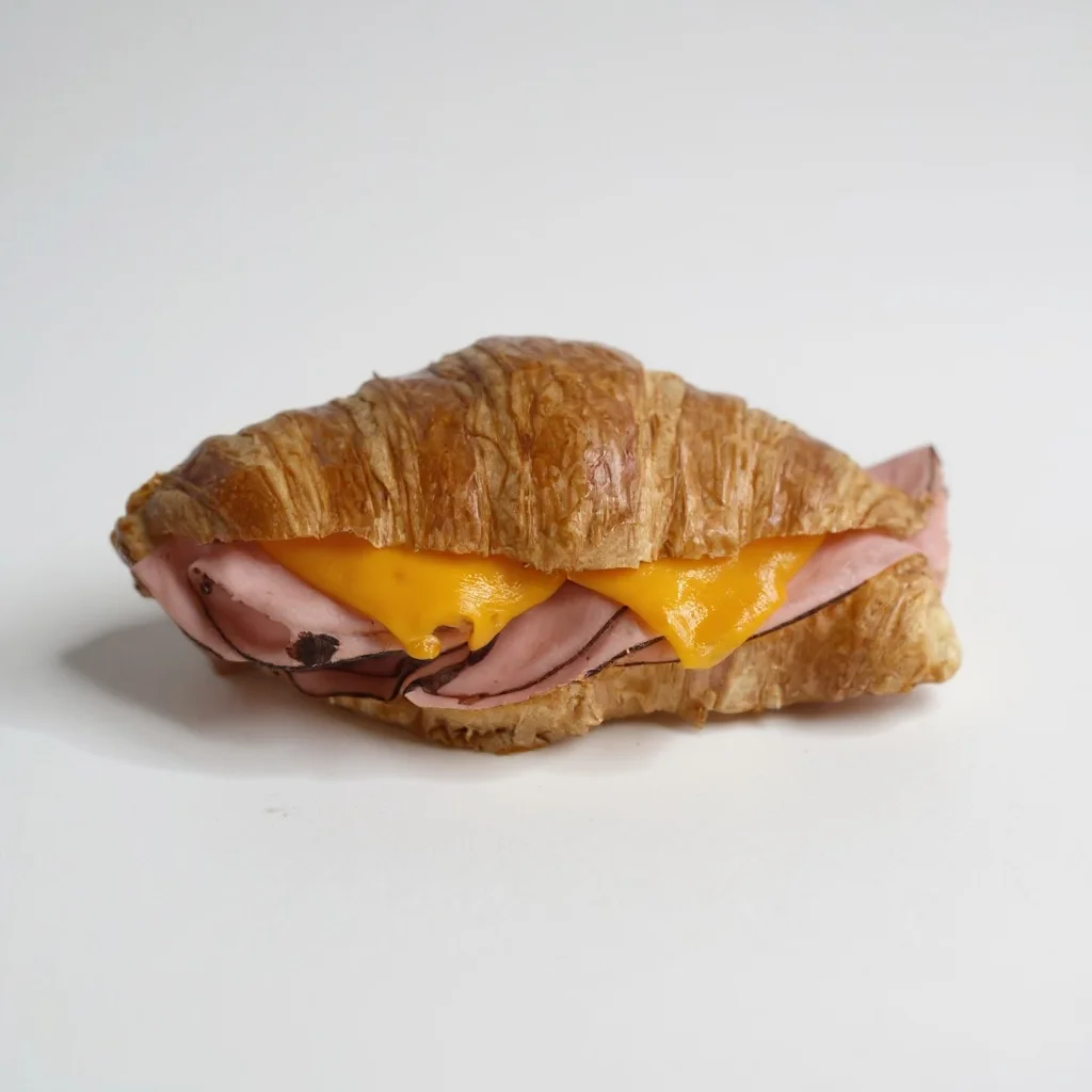 A croissant sandwich filled with layers of ham and melted cheddar cheese, arranged on a plain background.