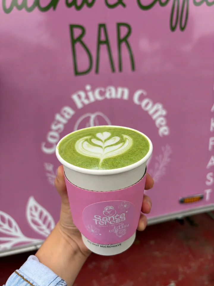 A cup of green beverage with a floral design on top, held against a pink background with coffee branding.