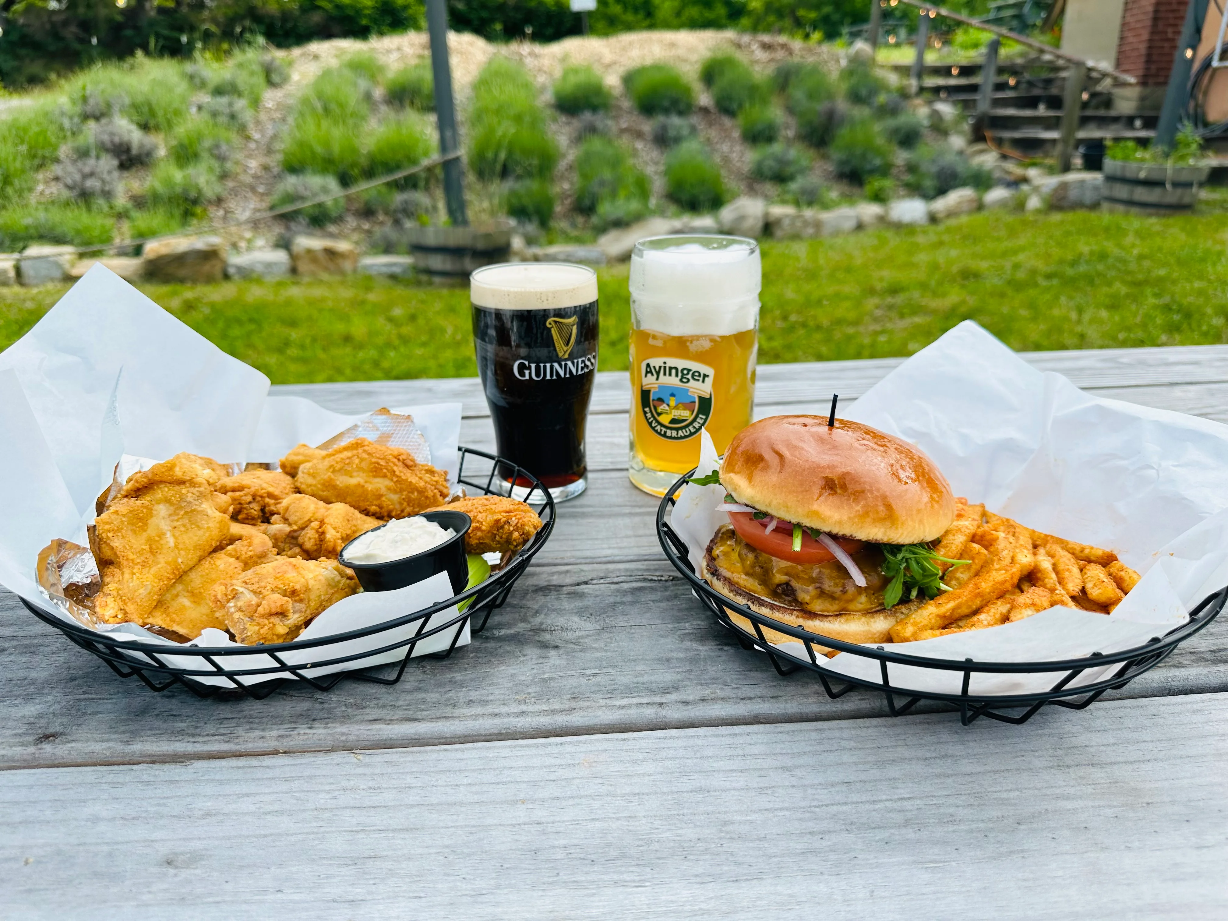 A delicious outdoor meal featuring fried chicken, a burger with toppings, and crispy fries, accompanied by two pints of beer on a wooden table.