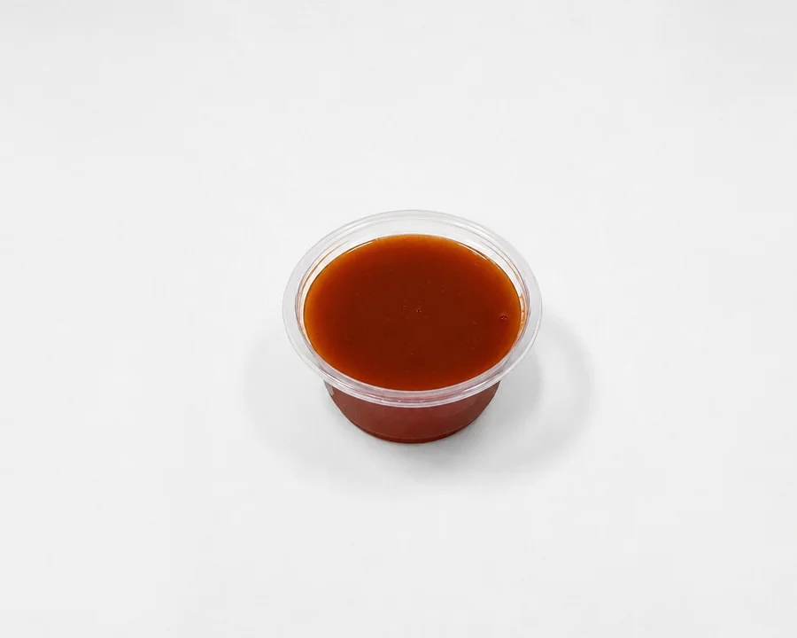 A small clear plastic container filled with a dark brown sauce, sitting on a white backdrop.