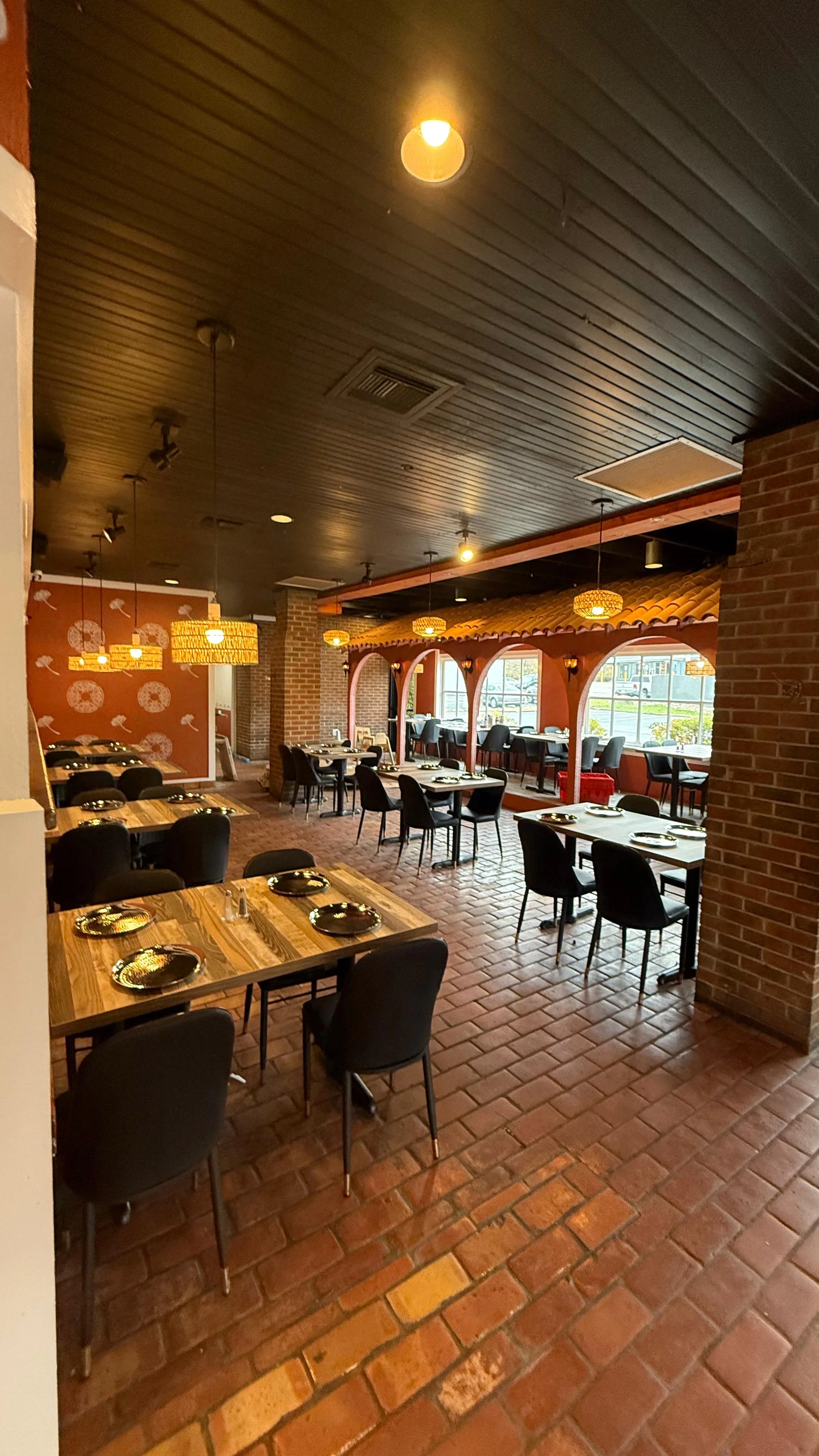 A cozy dining area inside a restaurant with wooden tables and black chairs. The decor includes hanging lights and warm tones, creating an inviting atmosphere.