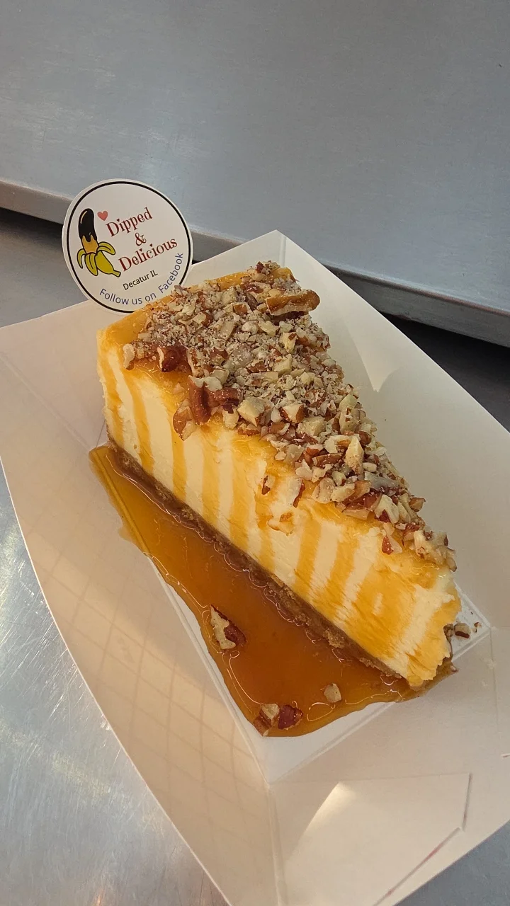 A slice of cheesecake topped with chopped nuts and a drizzle of syrup, served on a paper tray.