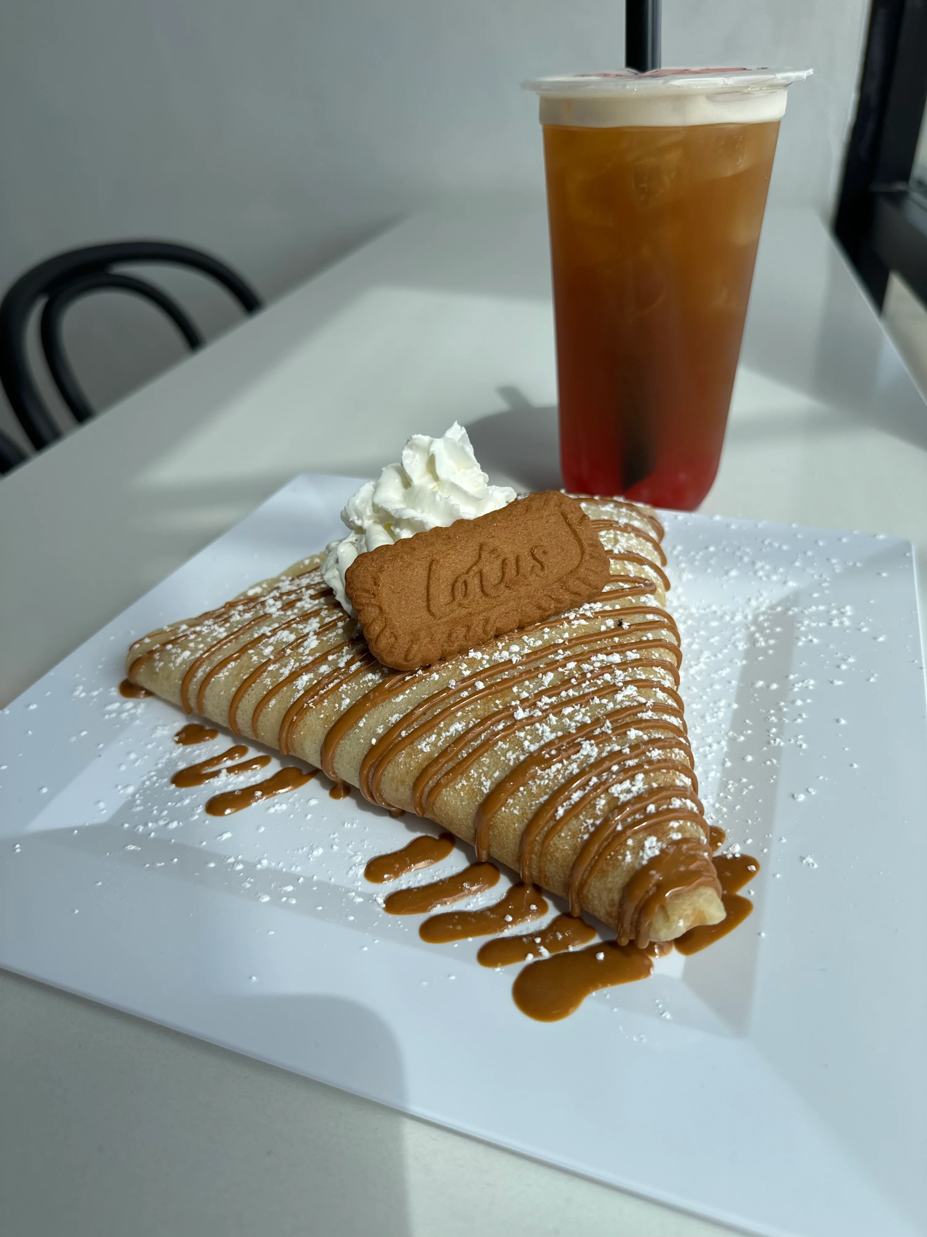 A triangular crepe topped with whipped cream, chocolate drizzle, crushed cookies, and colorful sprinkles, served on a plate.