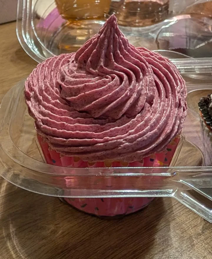 A pink frosted cupcake with a swirled decoration, placed in a transparent plastic container. The cupcake features colorful sprinkles at the bottom.