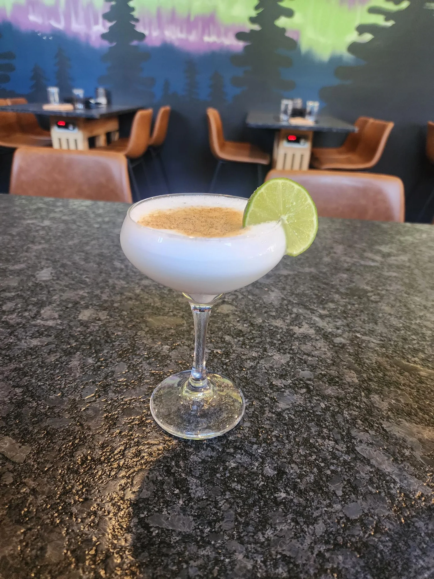 A cocktail served in a stemmed glass garnished with a lime slice, resting on a dark table with a colorful mural in the background.