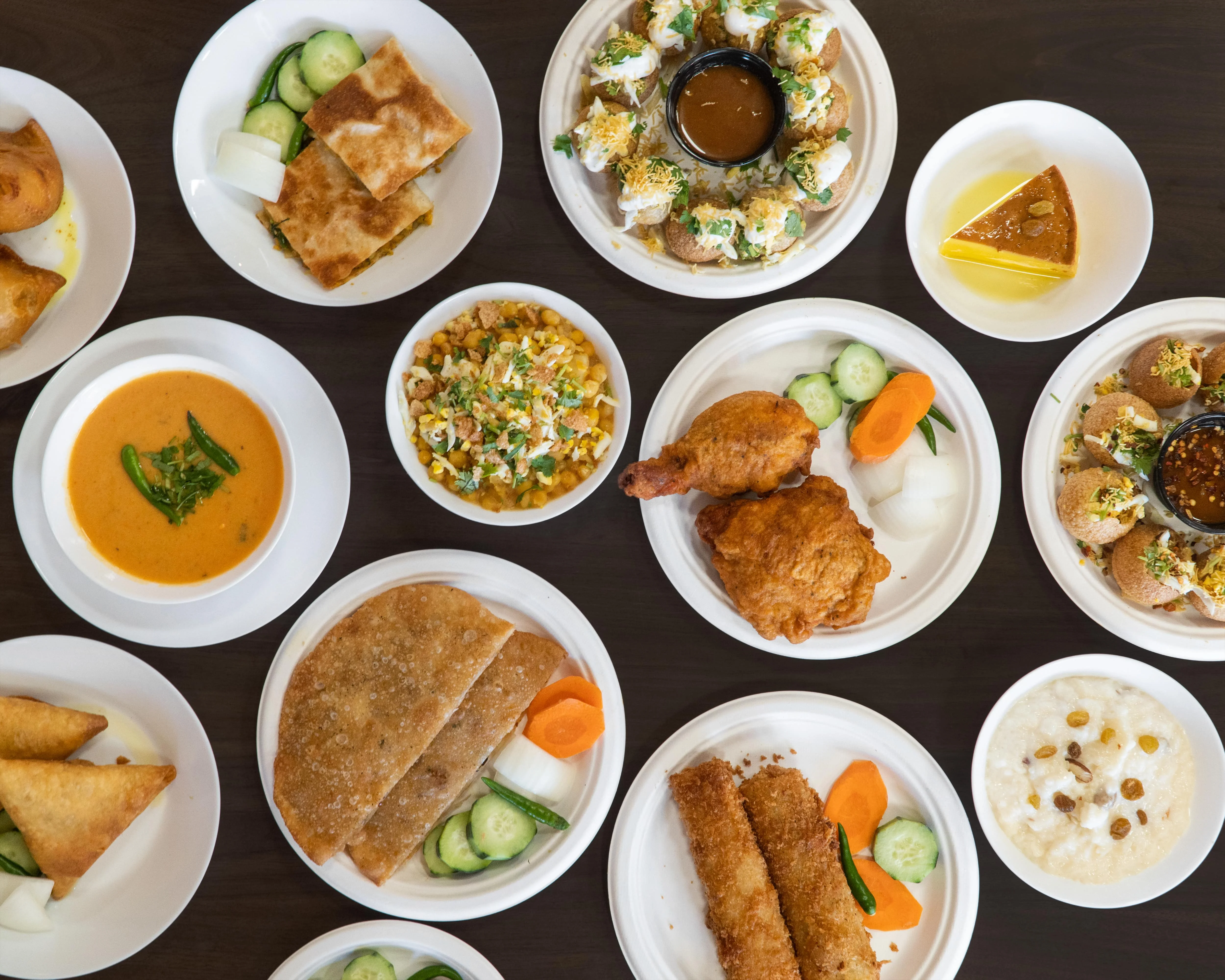 An assortment of Indian snacks and dishes including samosas, fried chicken, and a colorful dessert on white plates.