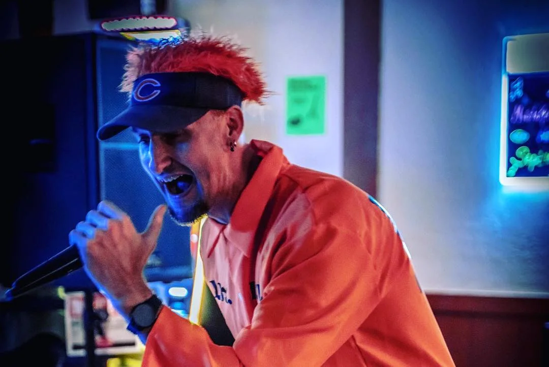 A performer in an orange outfit passionately singing into a microphone on stage, with colorful lights illuminating the background.