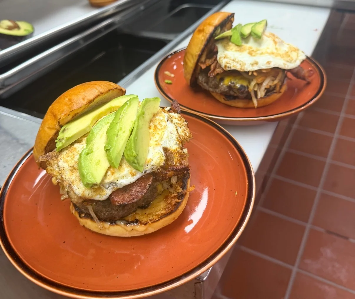 Two hearty burgers topped with avocado, fried eggs, and bacon on brown plates, showcasing a delicious meal.