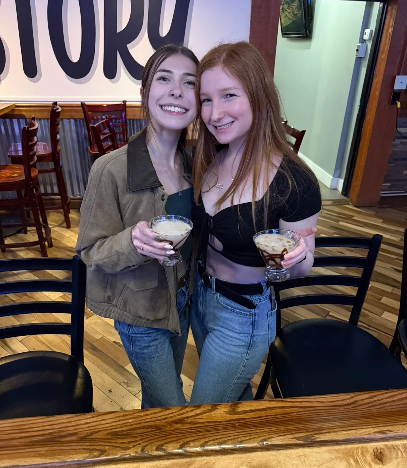 Two friends smiling and holding cocktails in a lively dining environment, showcasing a wood bar and casual seating.