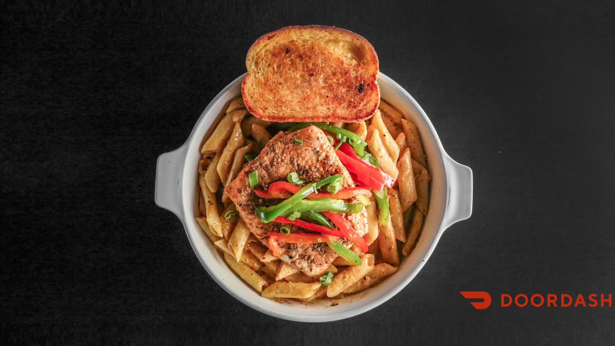 A bowl of pasta topped with grilled chicken, bell peppers, and green onions, accompanied by a slice of toasted bread.