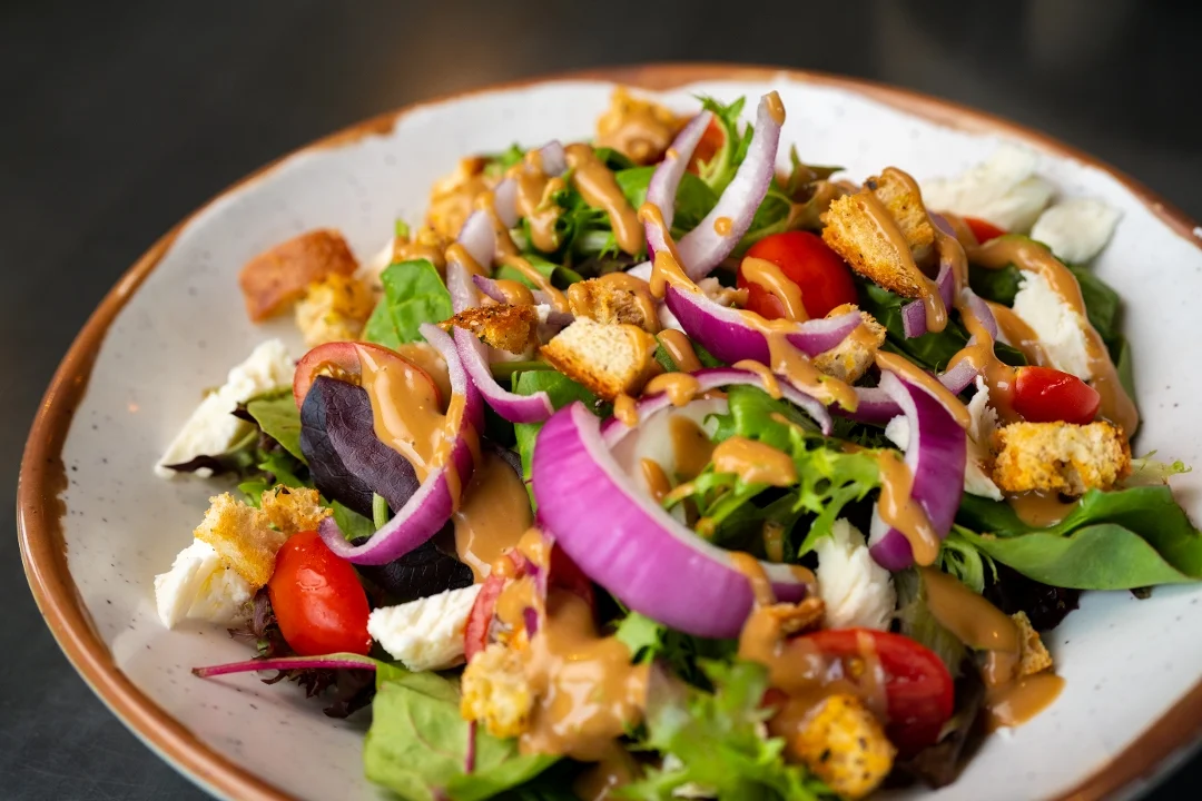 A vibrant salad composed of mixed greens, cherry tomatoes, croutons, red onions, and a drizzle of peanut dressing.