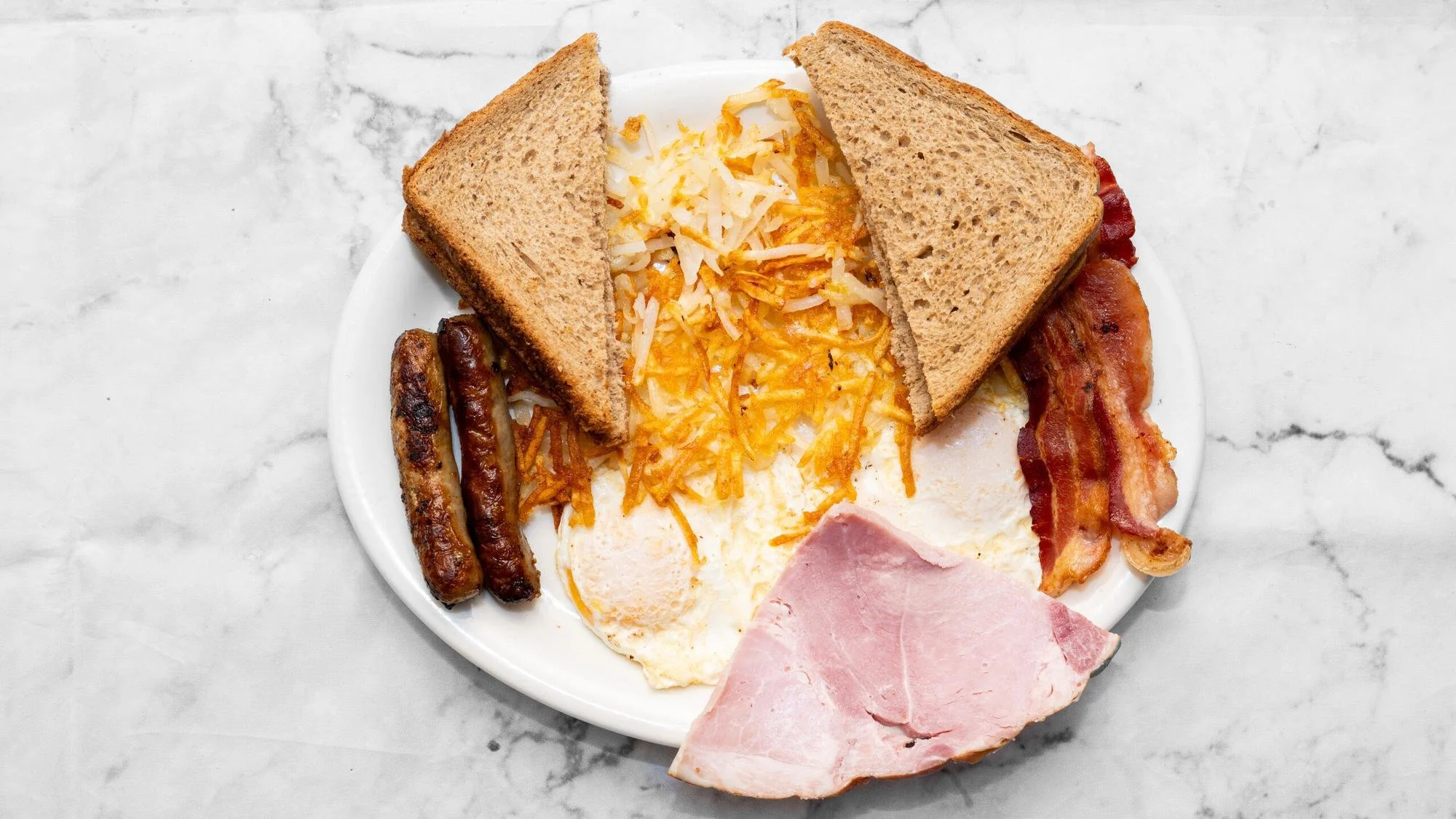 A hearty breakfast plate featuring two eggs, hash browns, two sausage links, strips of bacon, a slice of ham, and two pieces of whole grain toast.