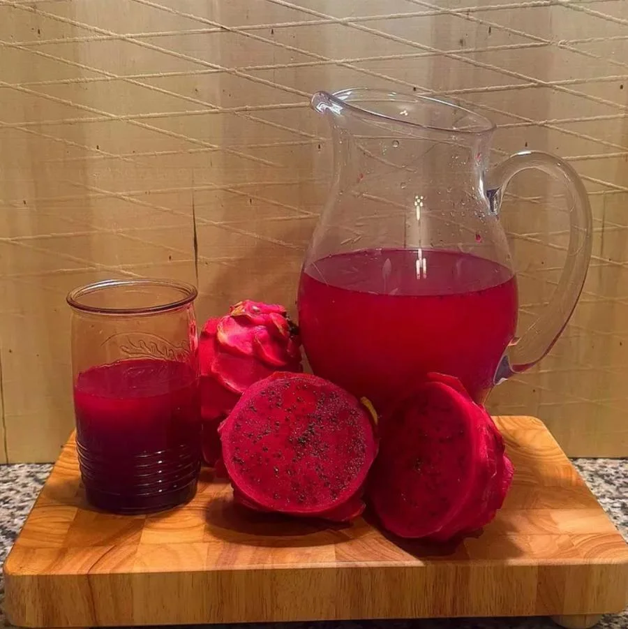 A pitcher of bright pink beverage next to a glass and dragon fruit slices on a wooden board.