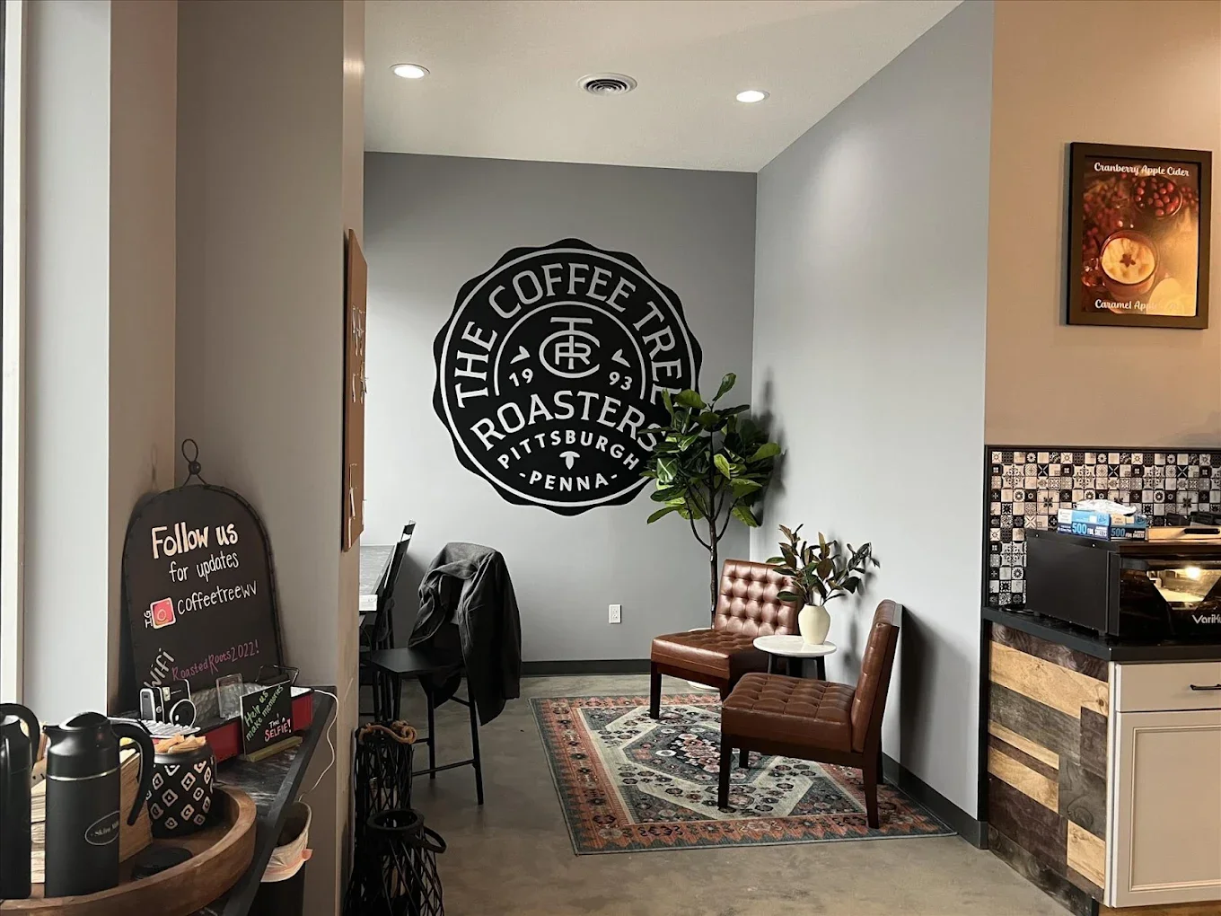 Cozy coffee shop interior with a large logo on the gray wall. Features two brown chairs, an indoor plant, and a small table on a patterned rug. Warm atmosphere.