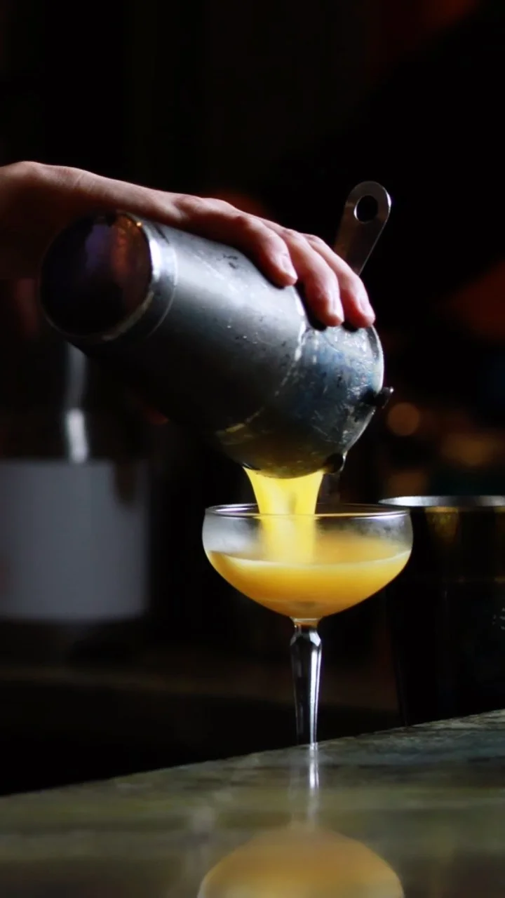 A close-up shot of a bartender's hand pouring a vibrant yellow cocktail from a chilled, frosted metal shaker into a delicate coupe glass, with a dark, blurred bar background.