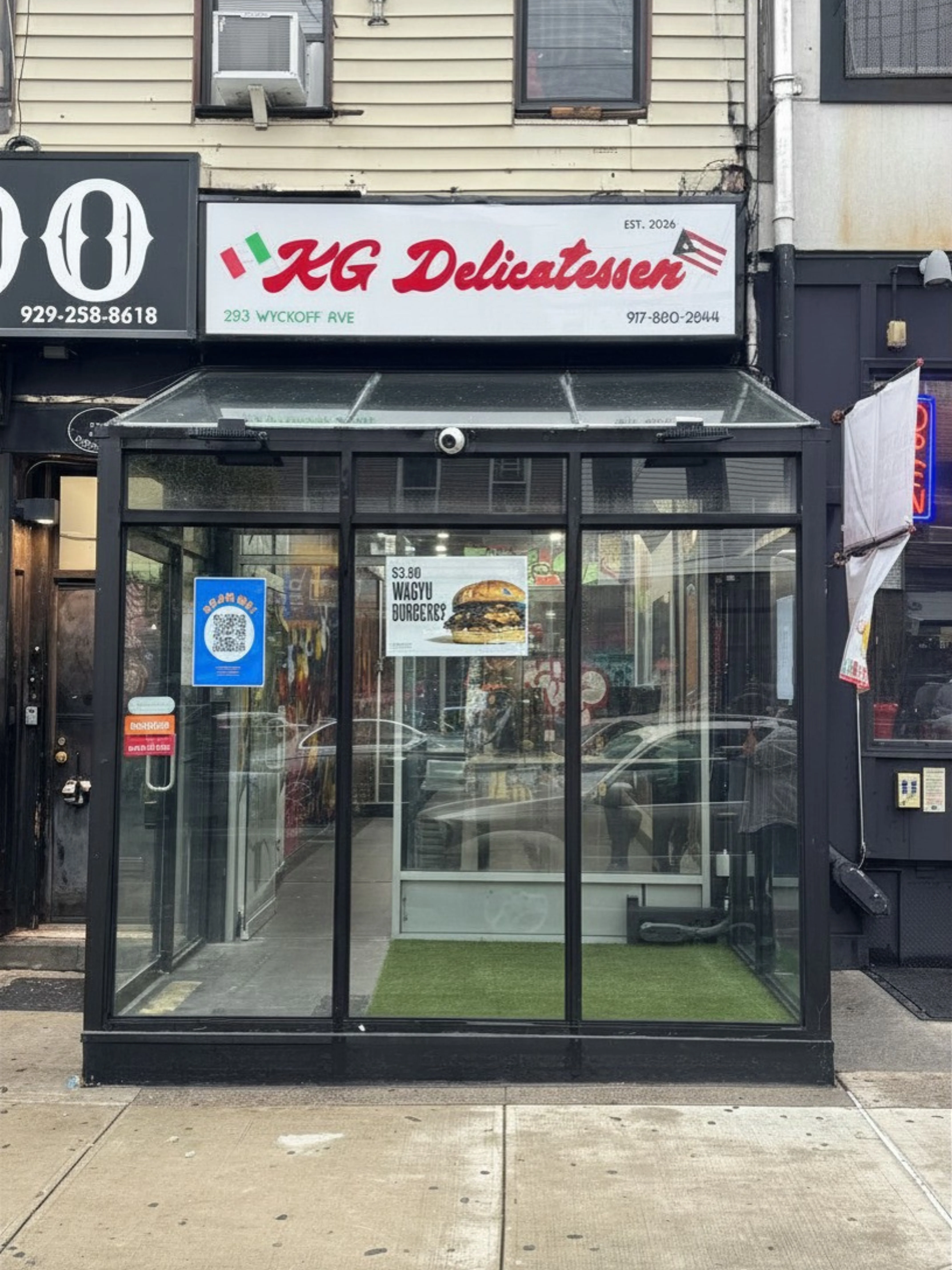 The exterior of a delicatessen with a prominent sign displaying its name and location. The storefront is glass-fronted, allowing a view of the interior.